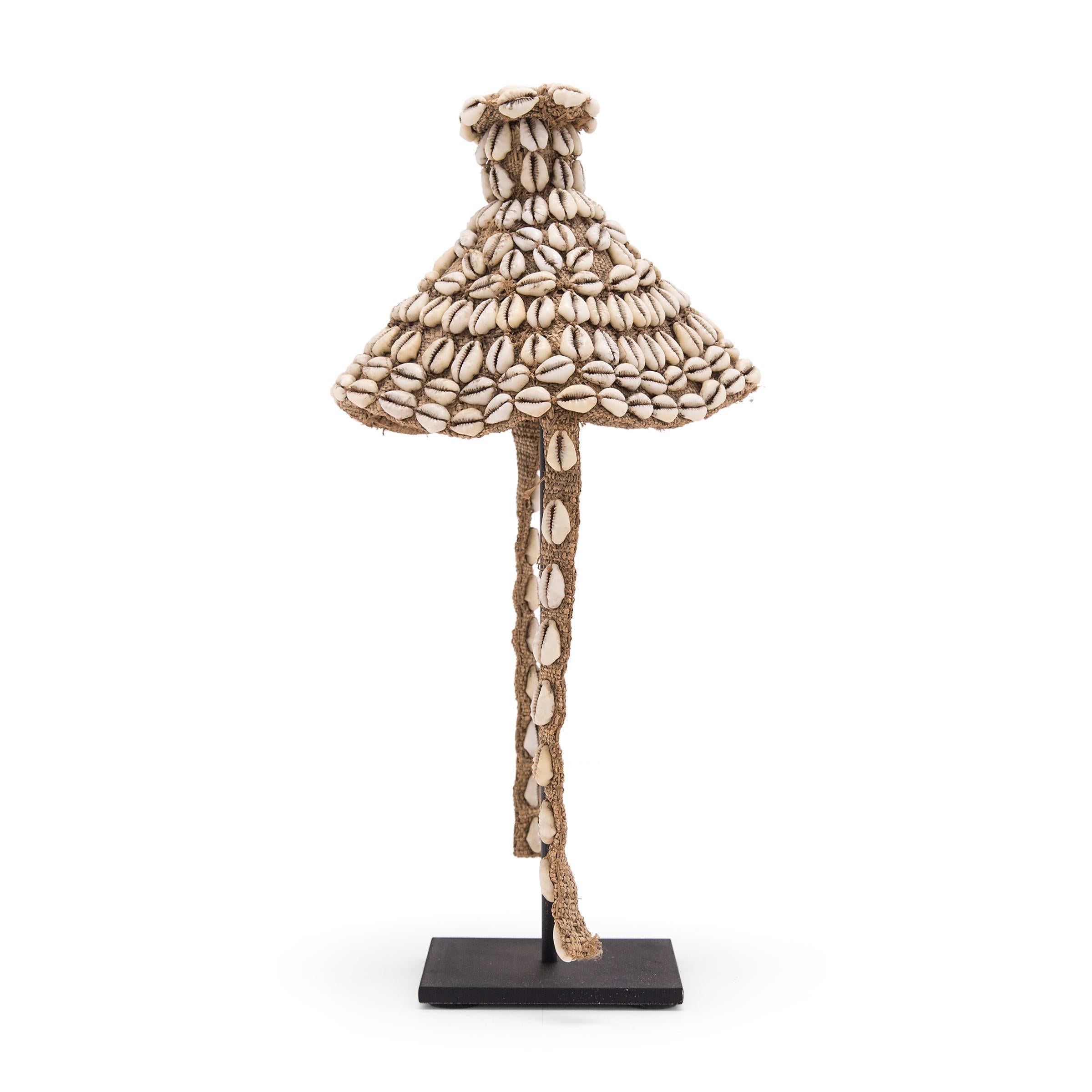This petite cone-shaped cap, known as laket, was created by an artisan of the Kuba peoples of the Democratic Republic of the Congo. The base is made of a simple woven raffia fabric, which was then embellished with a pattern of cowrie shells. Secured