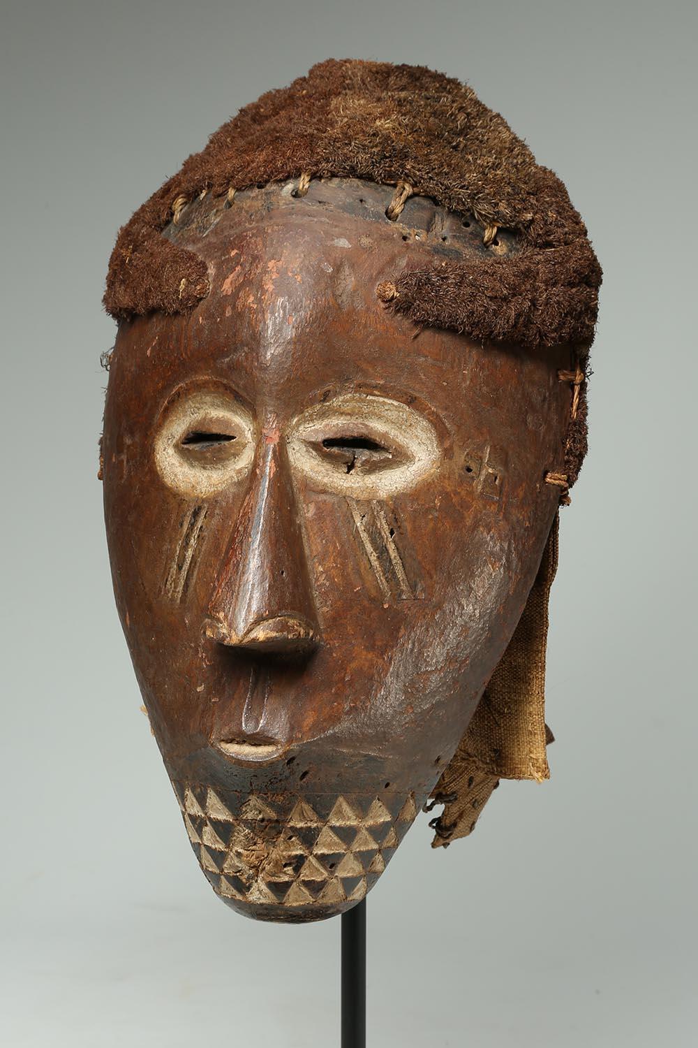 Early Kuba wood mask with traces of white and red pigments, geometric design on chin. Parts of raffia and cloth headdress attached. Woven raffia to simulate hair, early 20th century. On custom metal stand. Mask 13 x 7 x 8 inches. Old inventory label