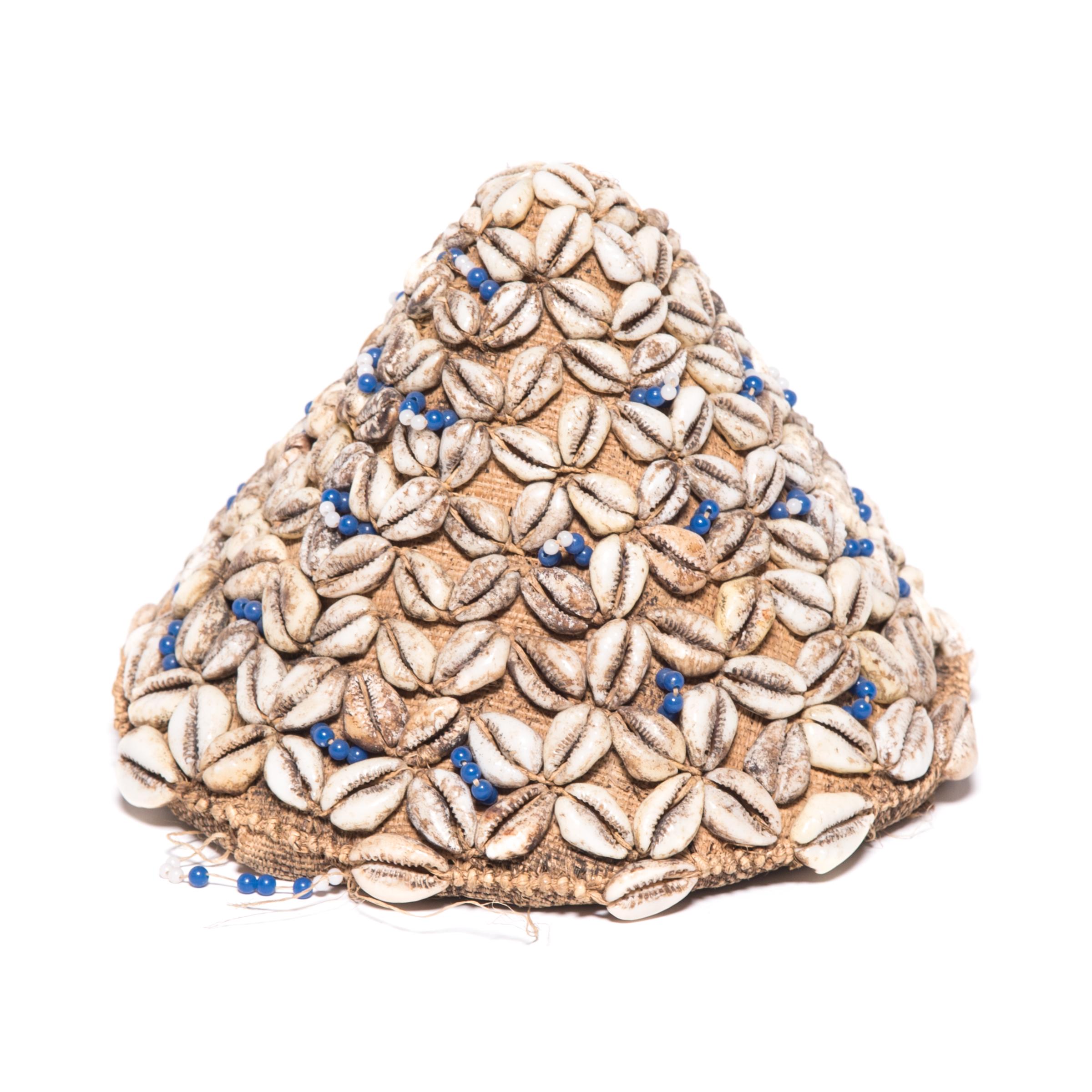This petite cone-shaped cap, known as laket, was created by an artisan of the Kuba peoples of the Democratic Republic of the Congo. The base is made of a simple woven raffia fabric, which was then patterned with cowrie shells and dotted with blue