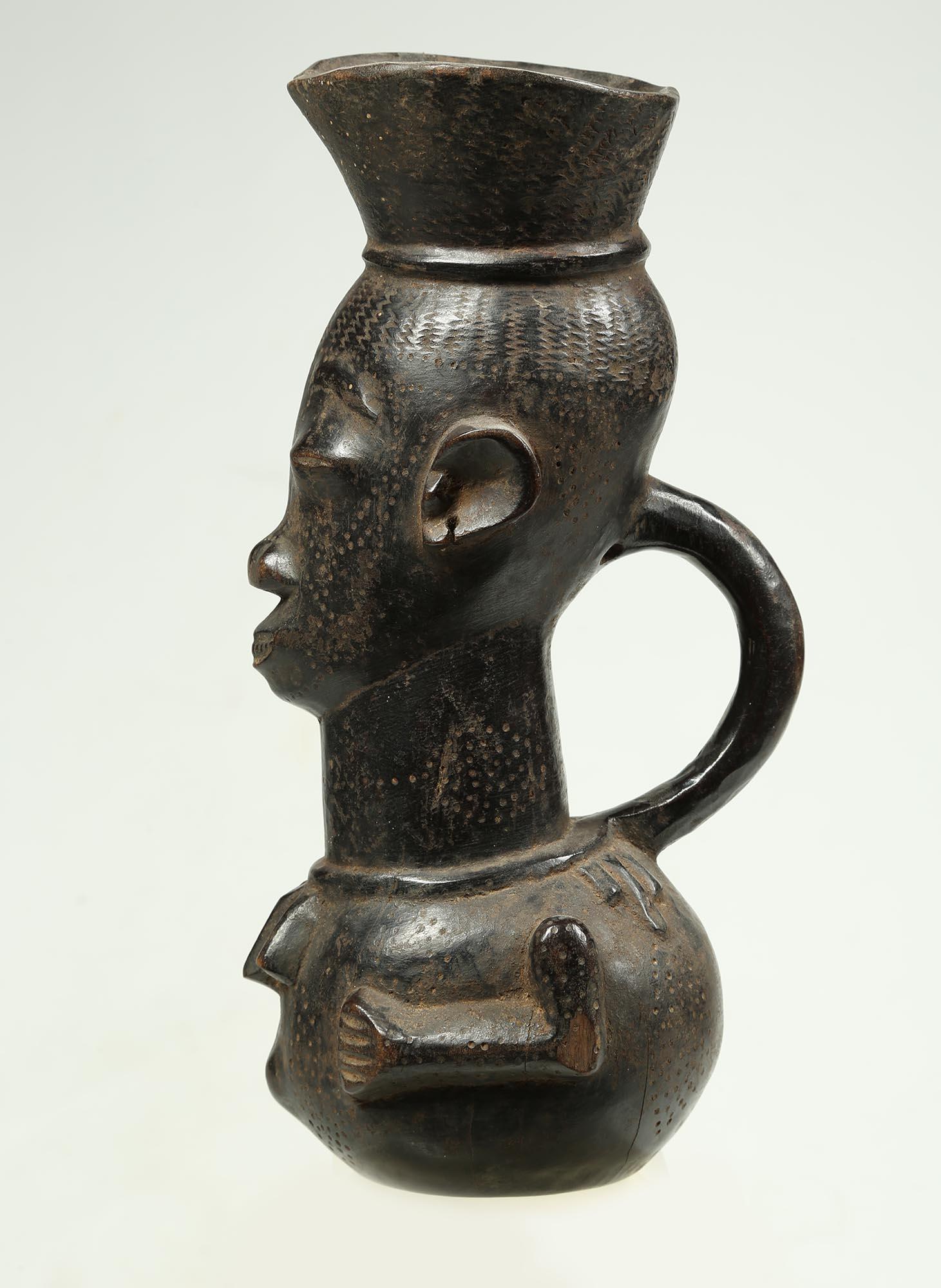 A tall Kuba palm wine cup of carved wood with large face and stylized body with hands of chest, handle and spouted top. 12 1/2 inches high, old stable repair to lip. Expressive face with wonderful expression. Kuba cups were items of high prestige