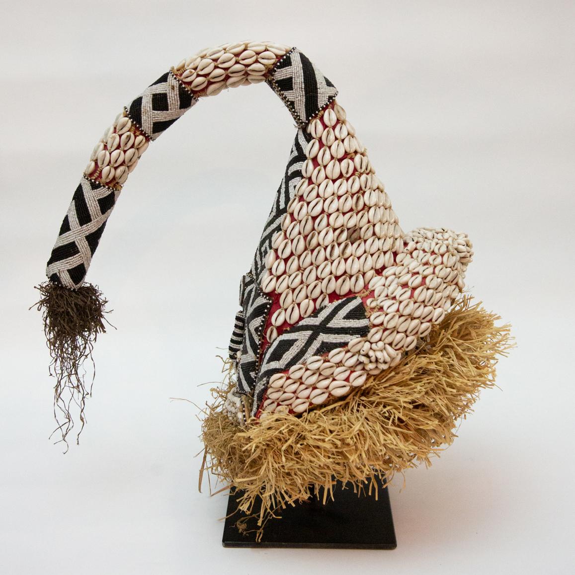 Elaborately trimmed with shell and hair decoration. Shell eyes. The surface is highly decorative, composed of intricately beaded geometric designs and cowrie shells. On red cloth base and trimmed with cream and brown raffia.