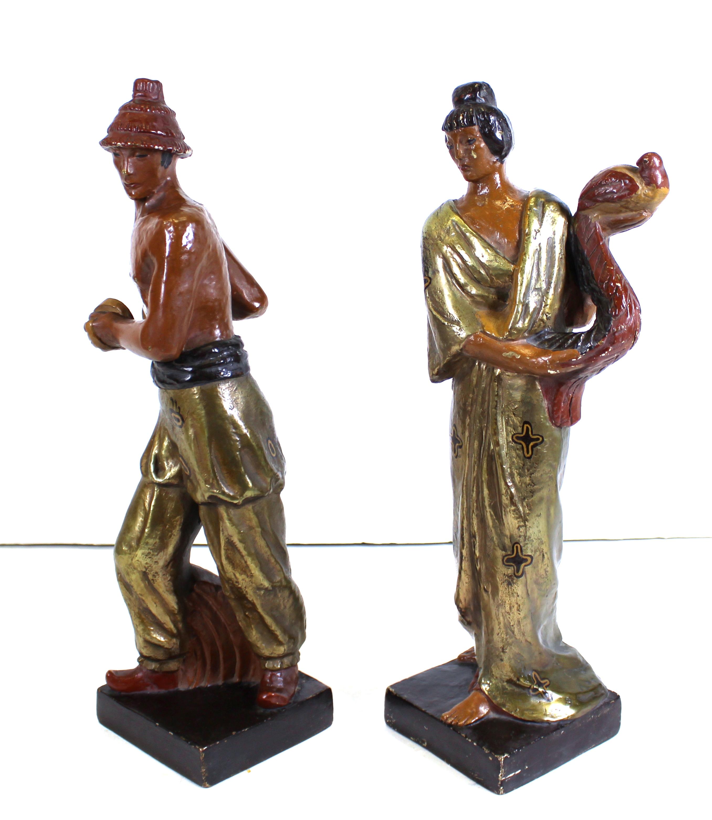 American Art Deco period pair of copper-clad terracotta sculptures of a couple in Japonesque style, a woman holding a peacock and a man with a hat holding musical instruments. The pair is silver and gold leafed and hand painted. Each piece is signed