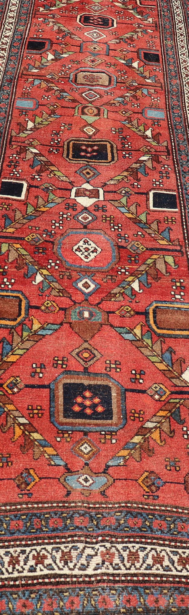 This early 20th century antique Kurdish runner features a triple-lined floral border, emboldening an ivory stripe between two blue edges. The field features a tribal, Sub-geometric pattern in a true red, accented by earthy tones in olive, ivory,