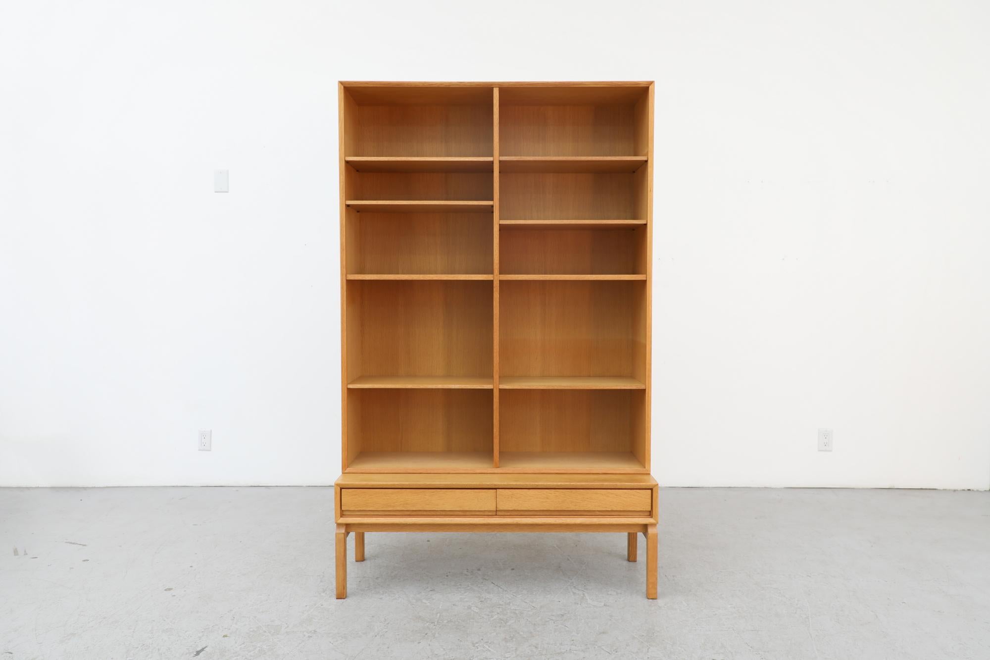 Mid-Century oak bookcase with lower double drawers. The bookcase is made of two separate pieces stacked together. The upper section are the bookshelves and the lower piece is wider with drawers. In original condition with visible wear, including sun