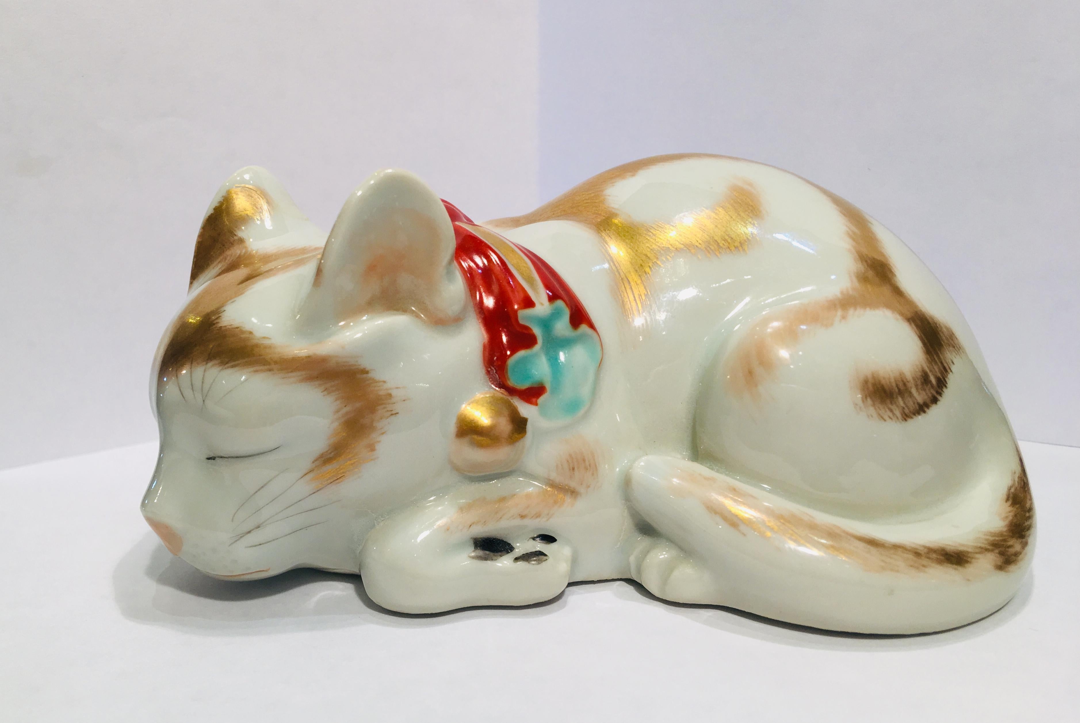 Very precious Kutani Japanese fine porcelain figurine of a curled up, peacefully sleeping little kitten is lavishly hand painted in 24-karat gold and features a large orange and gold bow with gold bells and blue accents.

From the Taisho period,