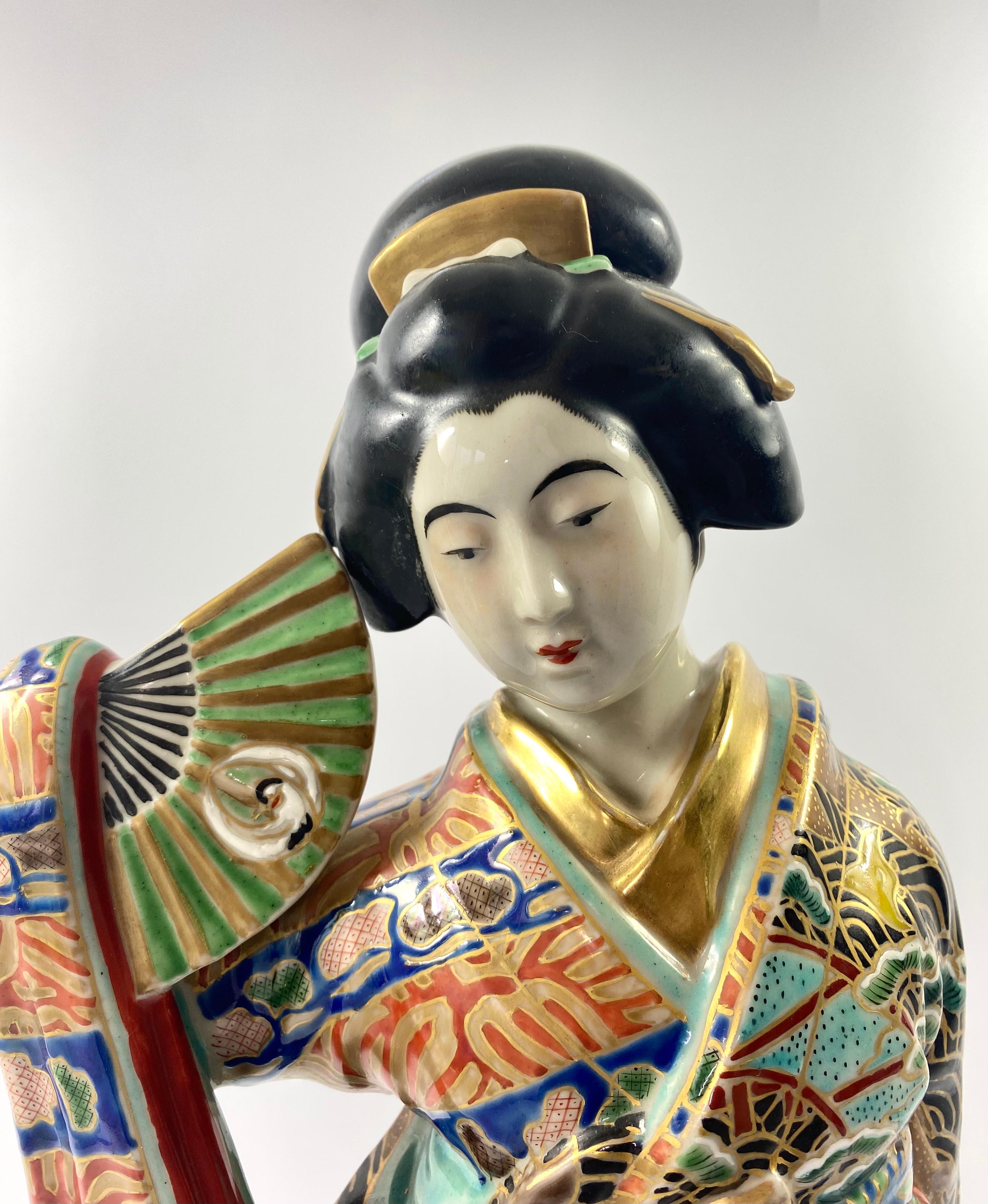 A large Kutani porcelain okimono of a Bijin, Japan, c. 1890, Meiji Petiod. The standing Bijin, dressed in an elaborately enamelled kimono, painted with a continuous river landscape of birds flying above snow covered pine trees. She holds a fan in
