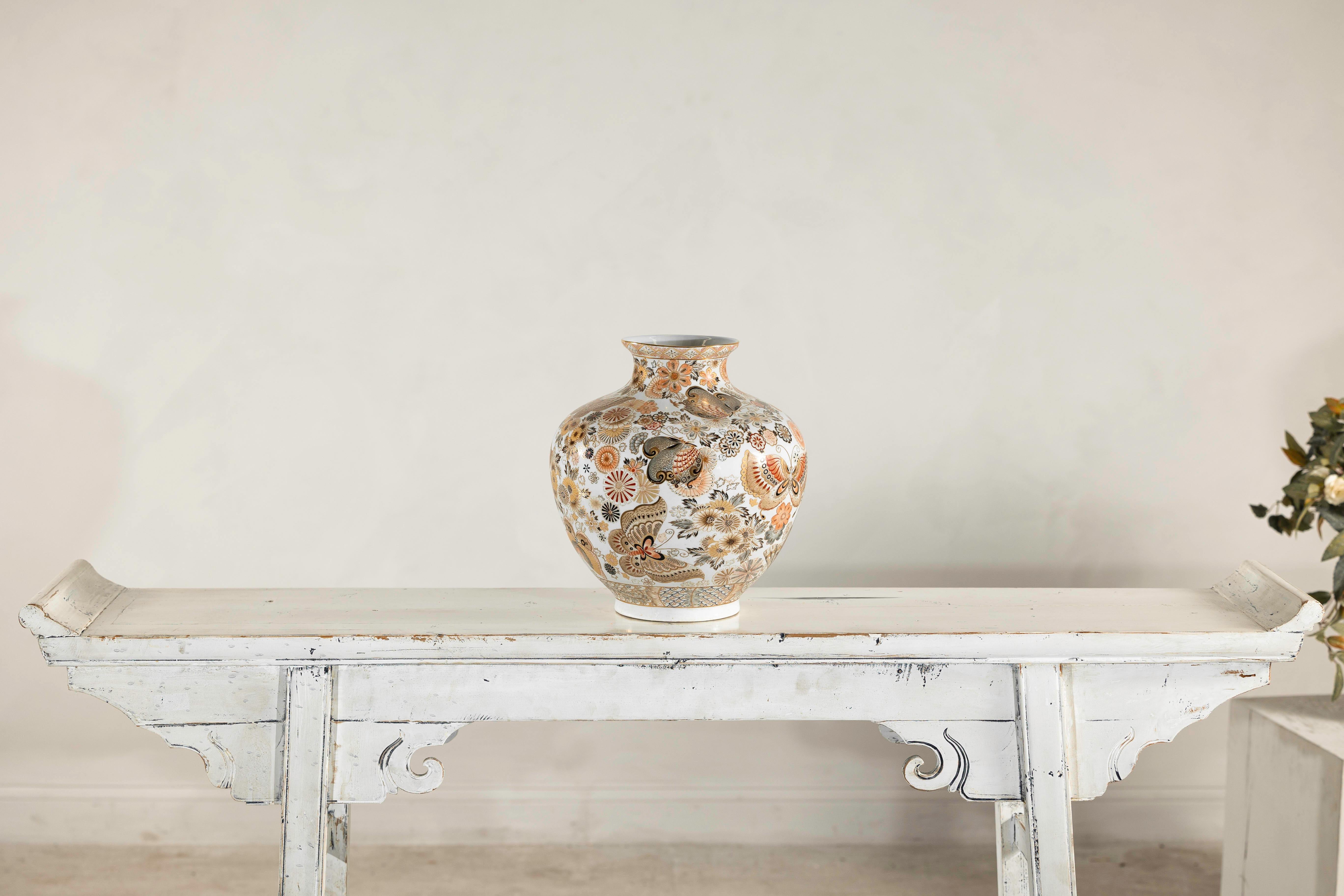 Un vaso in ceramica giapponese in stile Kutani riccamente decorato con un vivace disegno di farfalle e crisantemi su un fondo bianco. Questo pezzo d'epoca è una vetrina di intricati lavori di smalto, con decorazioni in sovrasmalto nei toni caldi del