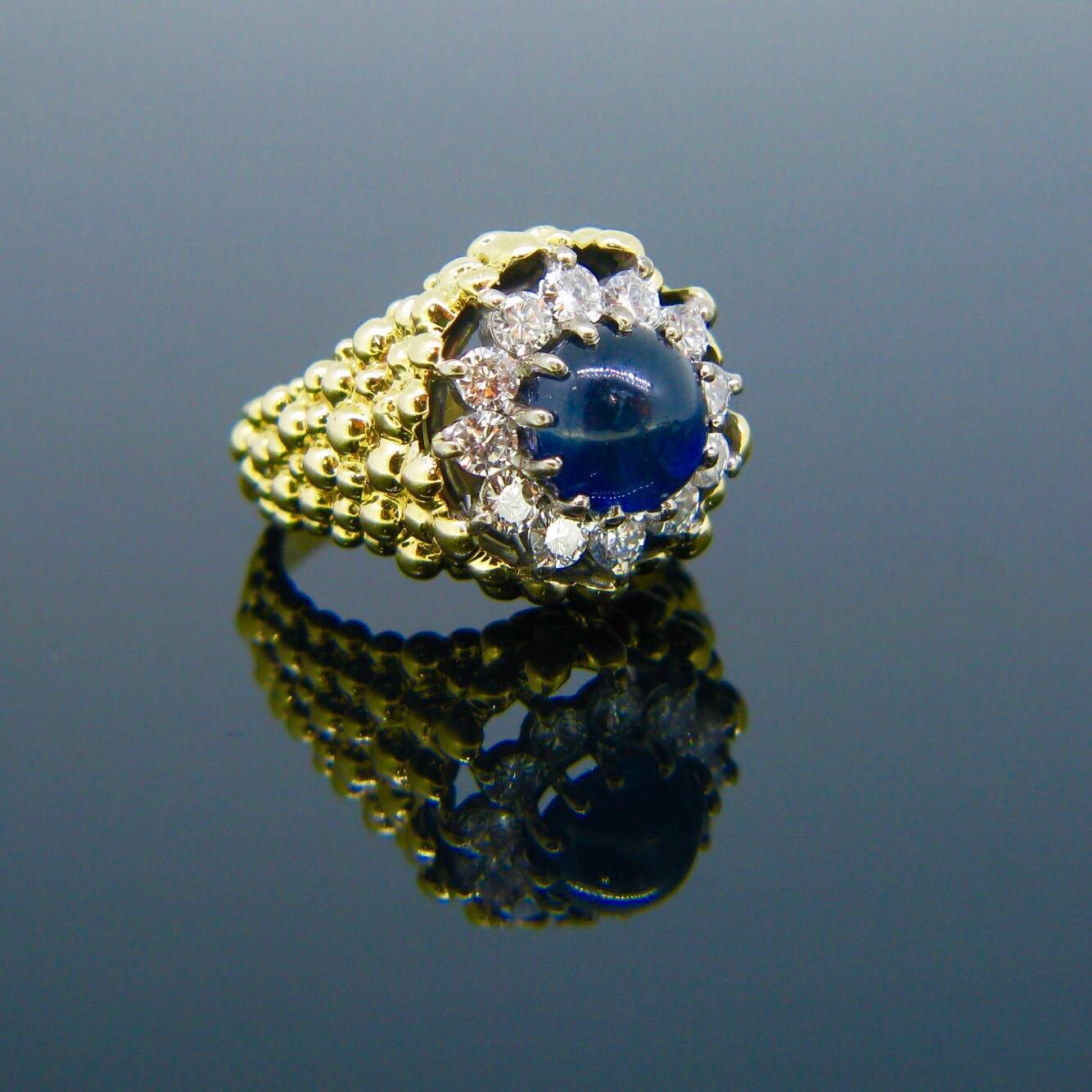 This impressive ring is signed Kutchinsky. It features a cabochon cut sapphire surrounded by 12 round brilliant cut diamonds. The ring is made in 18kt textured yellow gold giving the ring an unusual design.  It is signed inside the band