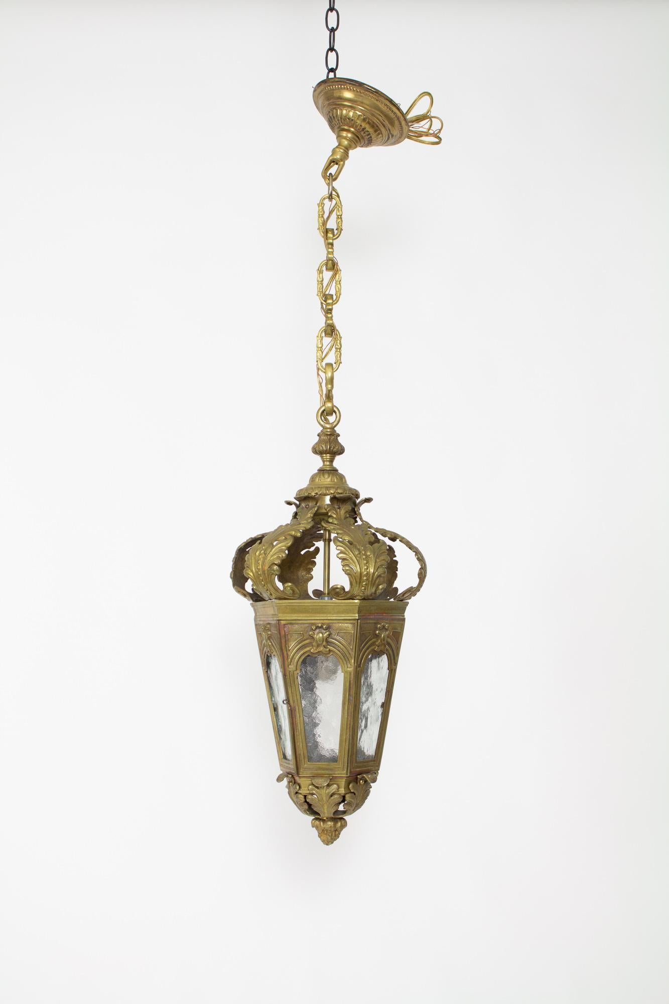 An ornate cast bronze lantern with acanthus leaf metalwork forming the top and bottom. There are six panes of textured glass. One of the six sides has a latching door for access to the single light bulb. This piece has been completely restored and