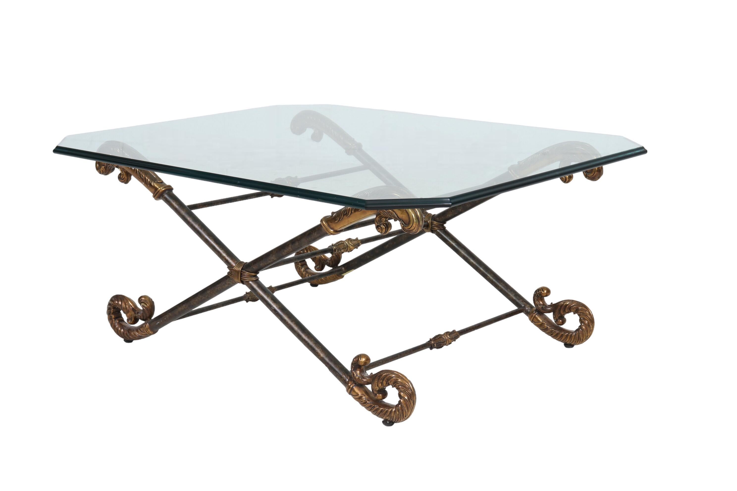 A brass and glass cocktail or coffee table made by La Barge. A rectangular glass tabletop with cut corners and a deep beveled edge rests on a double X shaped base, which is connected with five support bars. The feet are decorated with s-scrolls cast