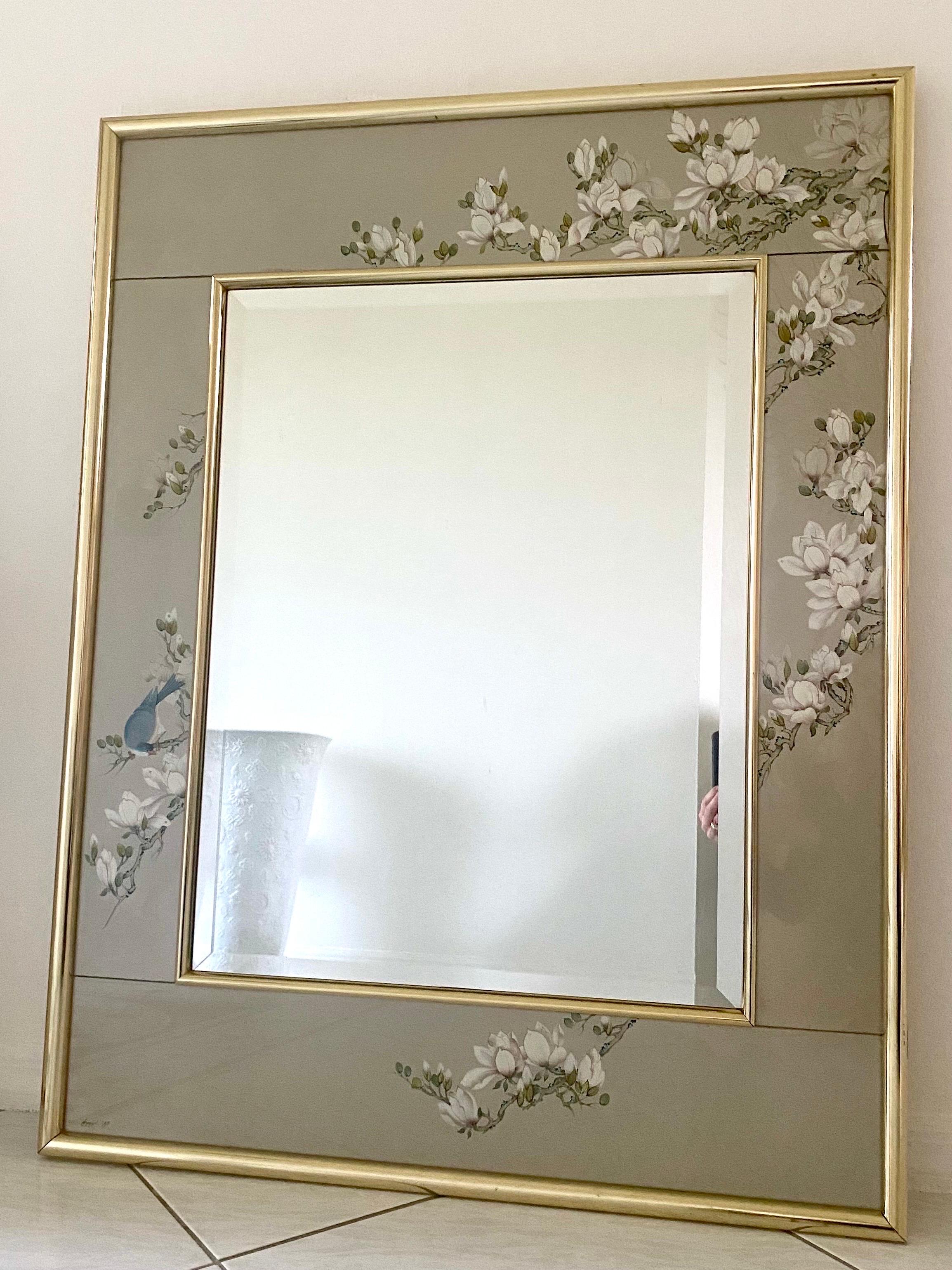 Artisan reverse hand painted glass mirror of flowering magnolia trees and bluebird, signed, dated 1989 with an outer frame in brass. The motif has exquisite details. The mirror is beveled. The mirror is ready to be hung and has its original La Barge