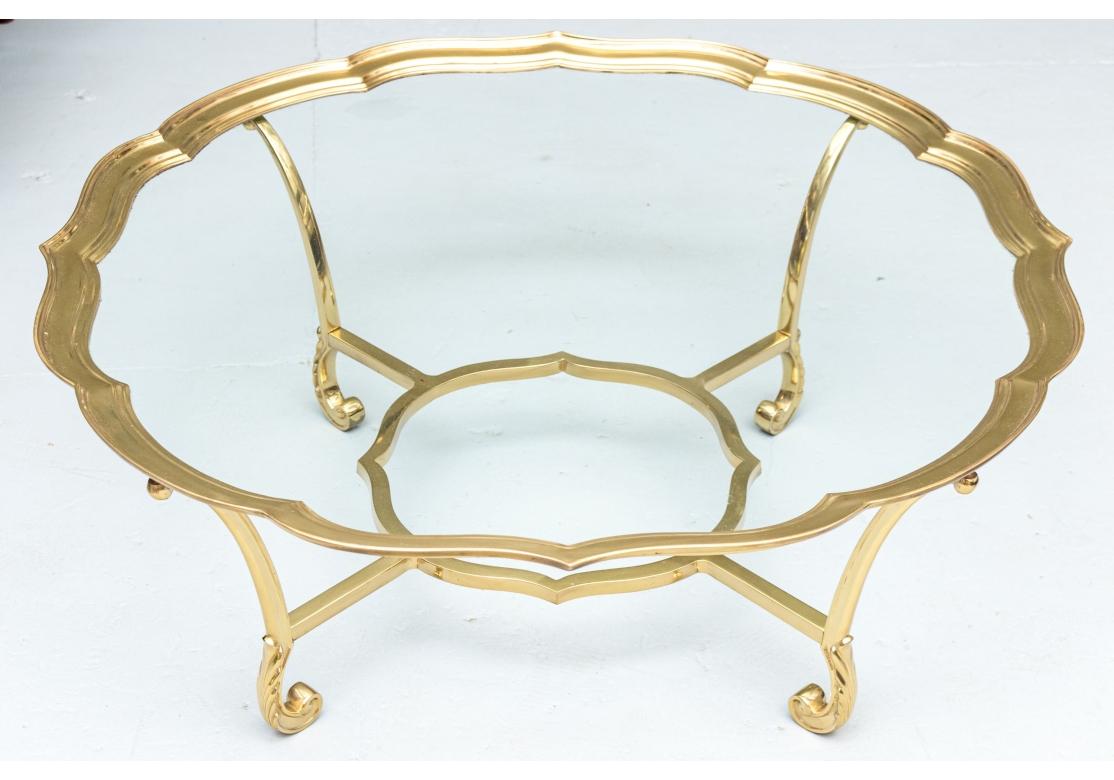 Hollywood Regency style solid brass coffee table by La Barge, featuring a large scalloped tray top inset with glass and tiered edges. The top is supported with four legs emitting from a shaped center stretcher and resting upon four acanthus leaf