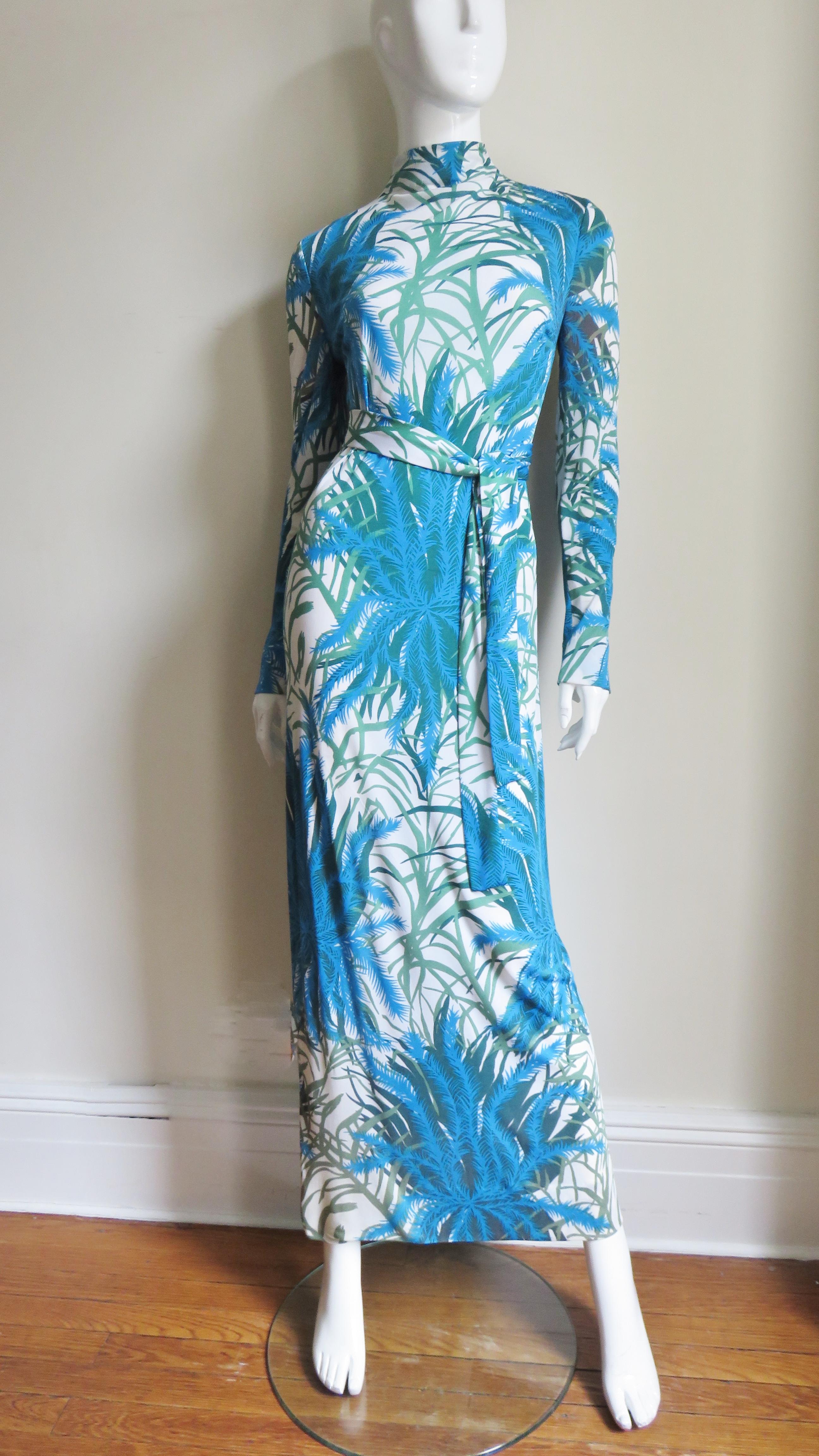 A maxi dress and over skirt in fine silk knit in a beautiful pattern of ferns and leaves in shades of green and blue on a white background from La Mendola. The dress is a long sheath with a stand up collar closing in the back with 3 dozen small self