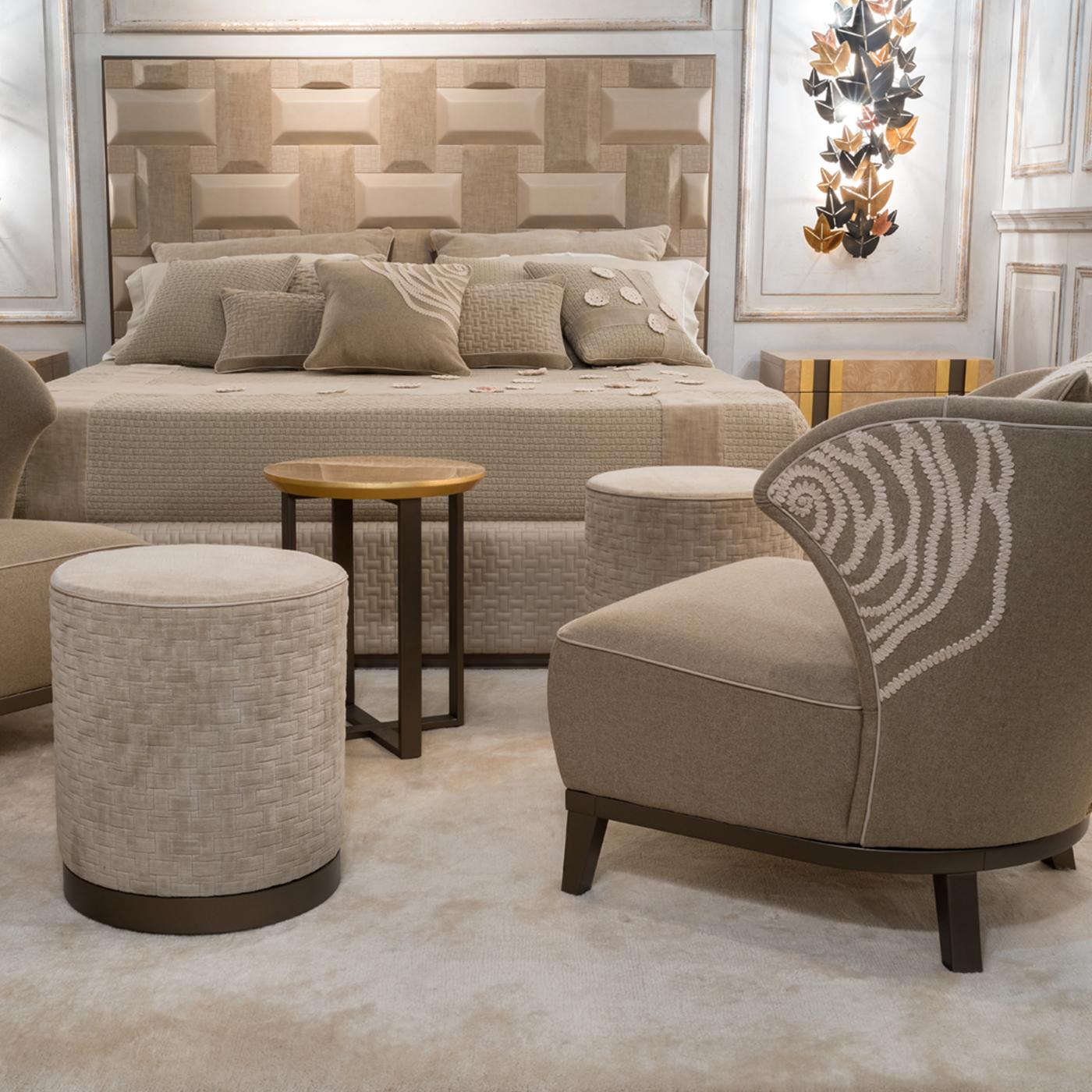 The Parigina Pouf by designers Marco and Giulio Mantellassi boasts a sleek cylindrical silhouette. Its padded seat is upholstered in versatile beige fabric, detailed with a textured motif for a woven effect. Supported by a sturdy bronzed steel base,