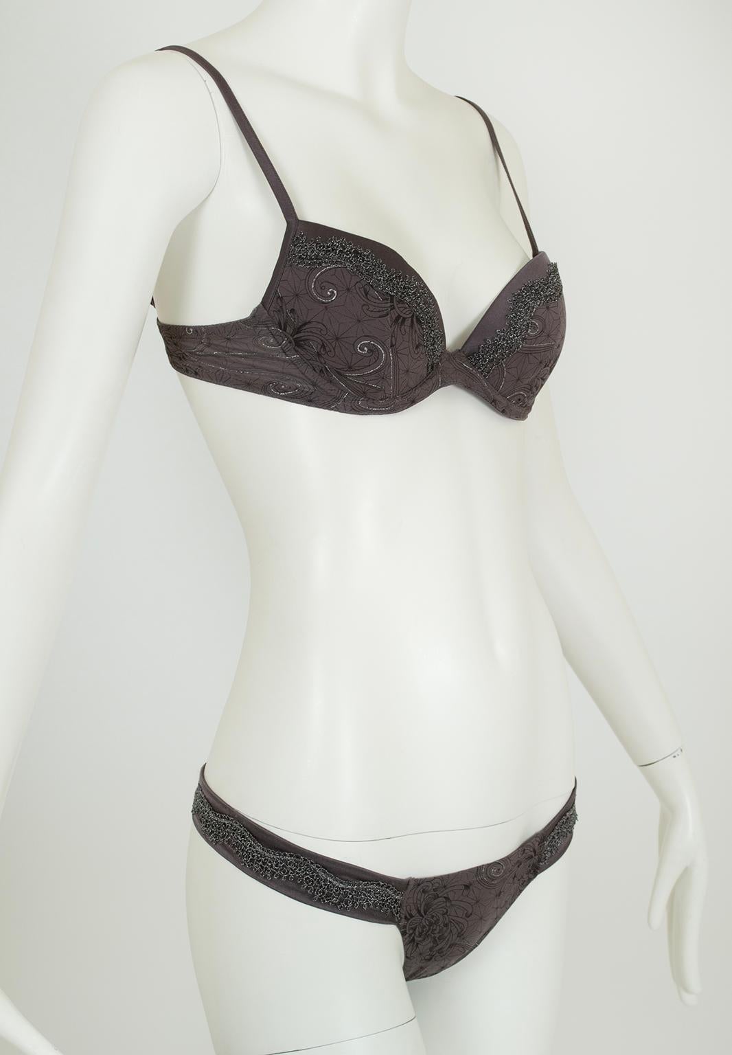 Reggiseno con ferretto e perizoma leggermente imbottito. Il reggiseno è caratterizzato da spalline regolabili e coppe in raso di seta color carbone; coppe e fascia in georgette di seta color argento metallizzato e impreziosite da pizzo color