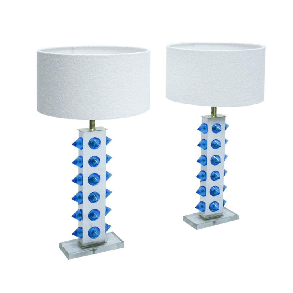 Ces lampes de table sophistiquées sont méticuleusement fabriquées en Italie, combinant le métal et le laiton avec d'exquises touches de verre bleu de Murano et d'élégants pieds en verre. Parés d'élégants abat-jour en tissu bouclé, ils témoignent