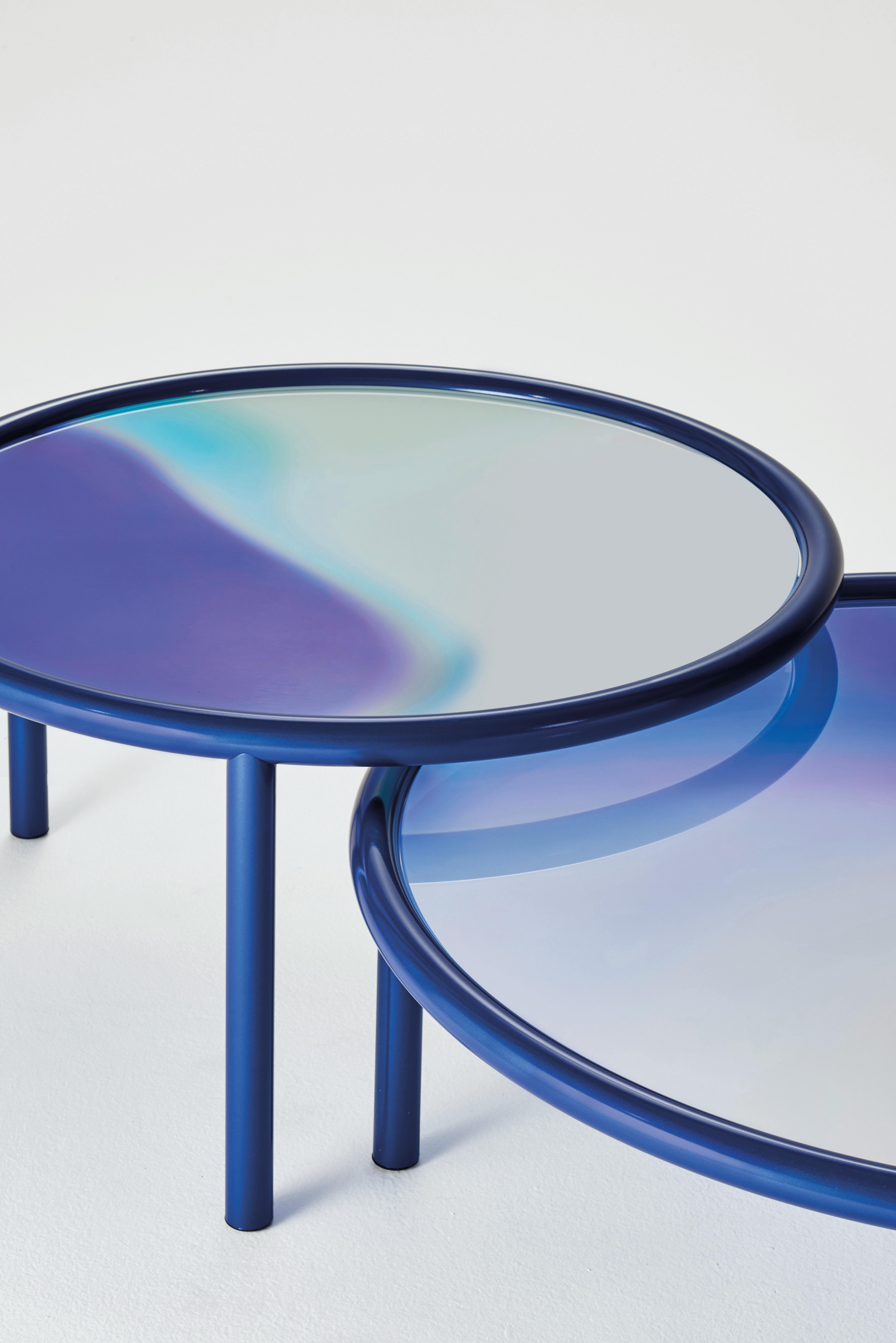 Low round tables with tubular metal structure painted midnight blue or copper colour in a special metallic opaque finish. The top, in laminated glass, is a palette of colors on a mirrored background, blending and mixing with one another like gas or