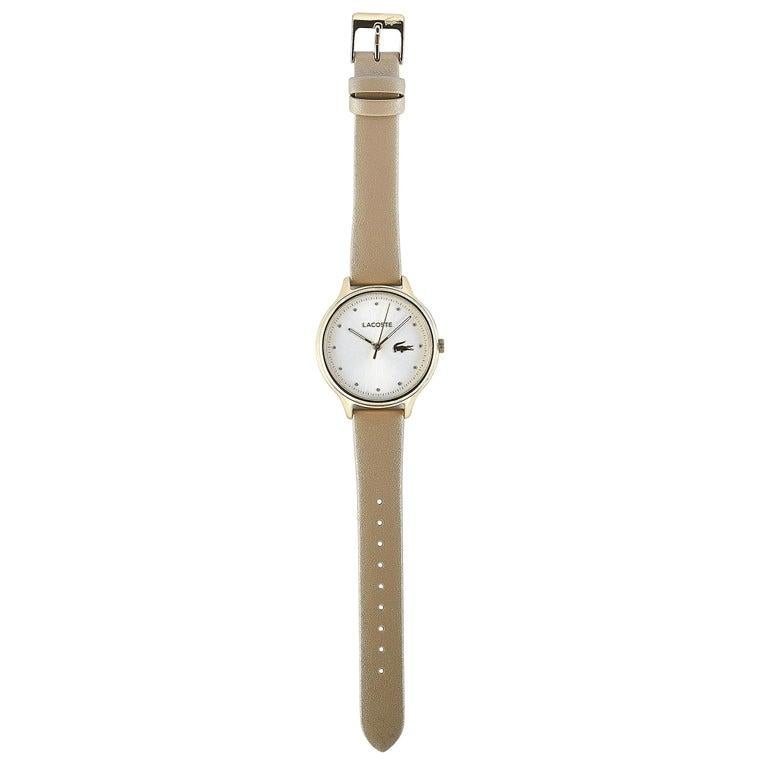 This is the Lacoste Constance watch, reference number 2001007. It is presented with a gold-tone stainless steel case that measures 38 mm in diameter. The case is water-resistant to 30 meters and mounted onto a pearly beige leather strap, secured on
