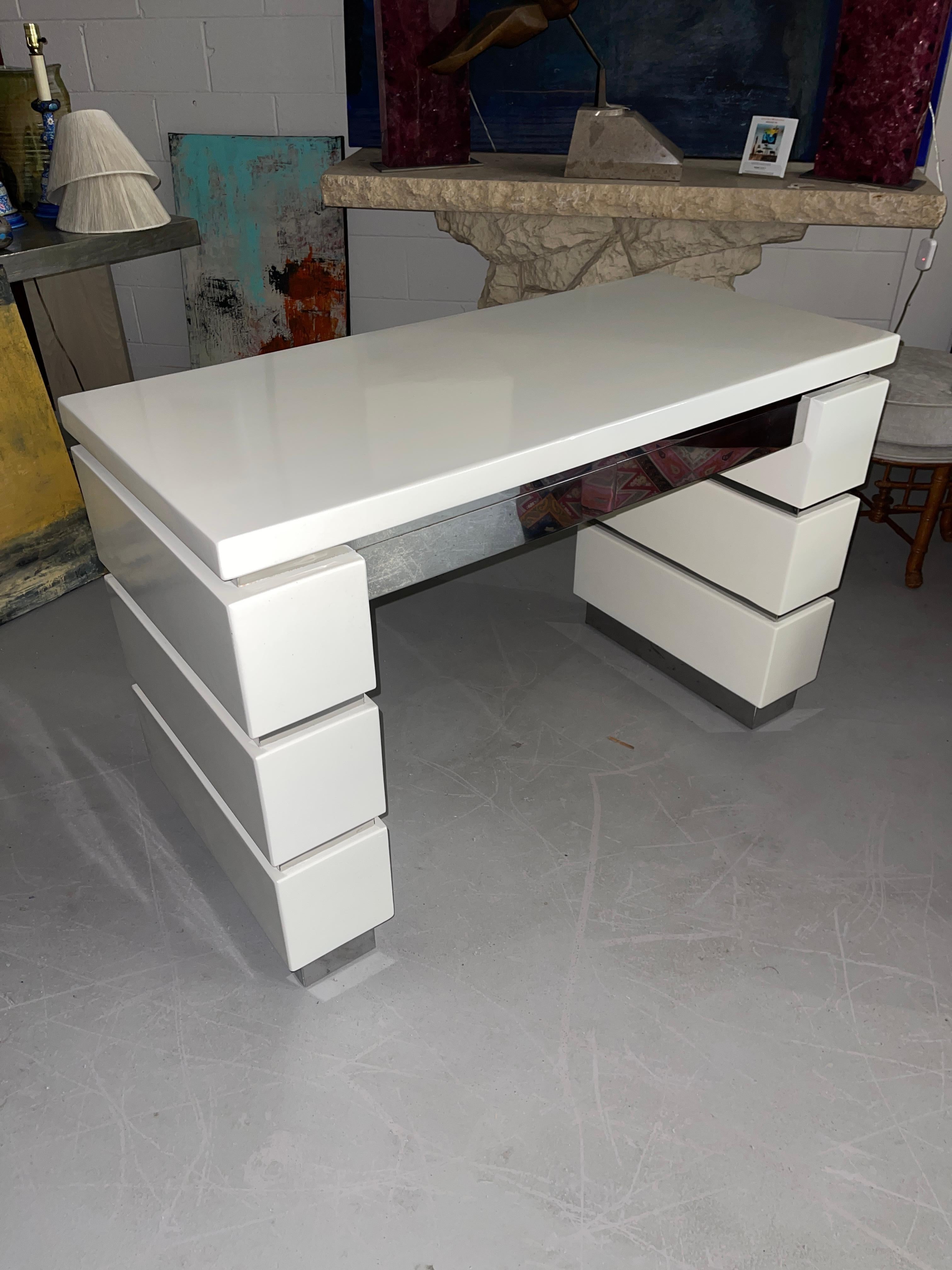 Una scrivania vintage laccata e cromata. Il colore è un bianco sporco/avorio. Ci sono due cassetti della scrivania con frontali cromati. I lati della scatola impilata hanno una fascia cromata. Il retro della scrivania è finito. Può stare liberamente