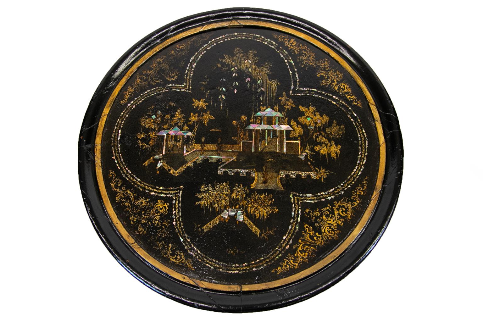 Lacquer occasional table has highlights of gilt arabesques that surround a quatrefoil center panel framed with mother of pearl inlay. This panel depicts a house with a stairway entrance and covered bridges with trees.