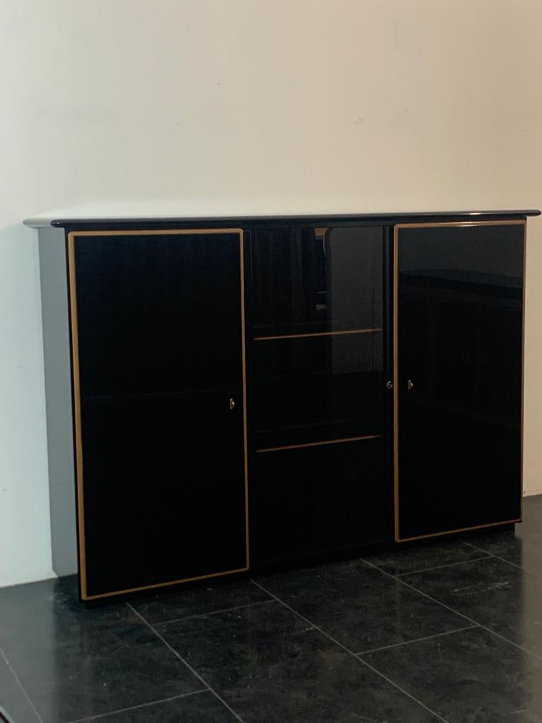Elegant sideboard characterised by two side doors and a central sliding glass. The cabinet is lacquered black, the central glass is smoked black. It features a precious layered wood profile that outlines the edges of the doors and shelves,