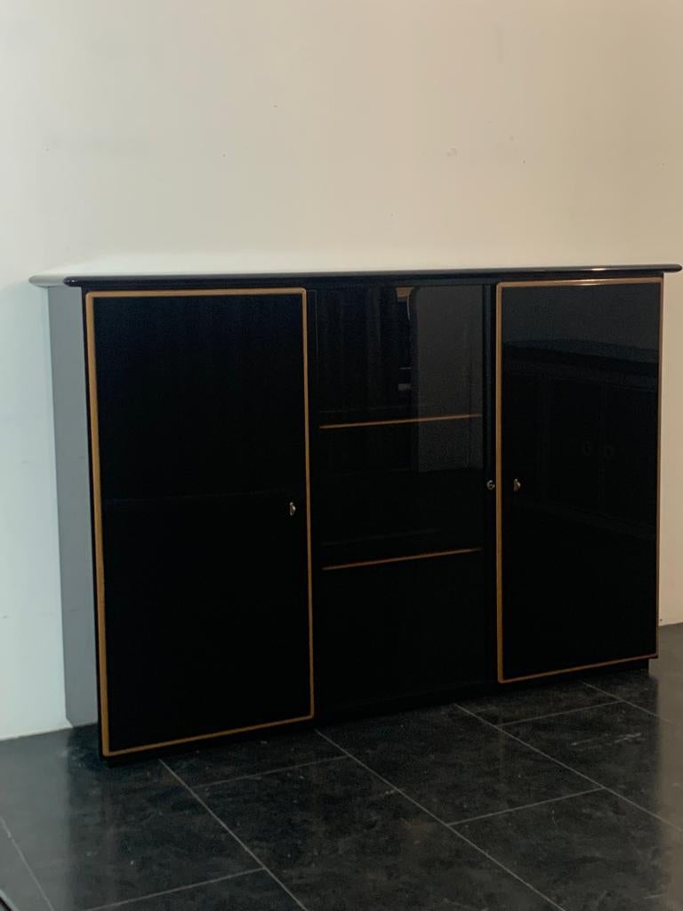 Elegant buffet caractérisé par deux portes latérales et une vitre centrale coulissante. Le meuble est laqué noir, le verre central est noir fumé. Il présente un précieux profil de bois stratifié qui souligne les bords des portes et des étagères,