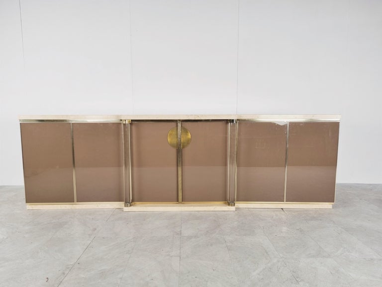 Luxurious seventies glamour sideboard by Maison jansen consitsing of glass and brown lacquered doors, brass finishes, lucite columns and travertine base and tops.

The use of different high quality materials makes this piece a real eye