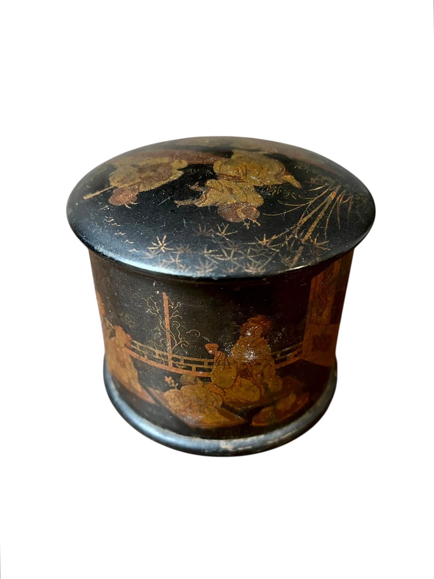 A charming cylindrical box with a domed lid made of black lacquered papier-mâché, known as japanning, is decorated throughout with chinoiserie scenes featuring small figures in a garden, a balustrade, bamboo, and rocks, executed with gold dusting