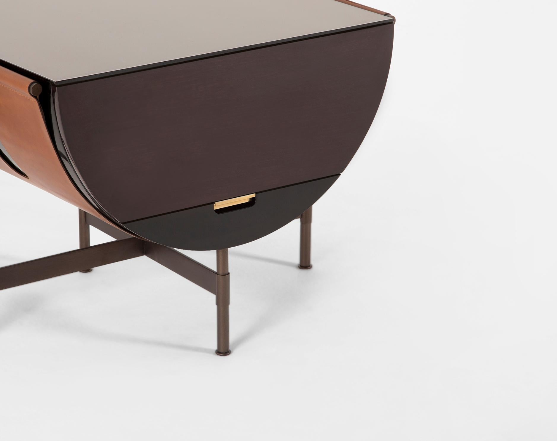 A hammock-like wide leather strap supports a half circle form and is lacquered black. The leather straps are wrapped around T-shaped steel arms. A brass arm extends to support the cabinet door when open. The Packsaddle Side Table is accompanied with