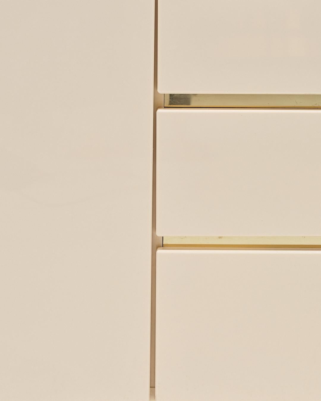 Buffet vintage des années 1970, en bois laqué beige avec éléments en laiton. Composé de deux portes battantes ouvrant sur des étagères et de quatre tiroirs de rangement, avec base et rangements cerclés de laiton. Bon état général, dans l’esprit des