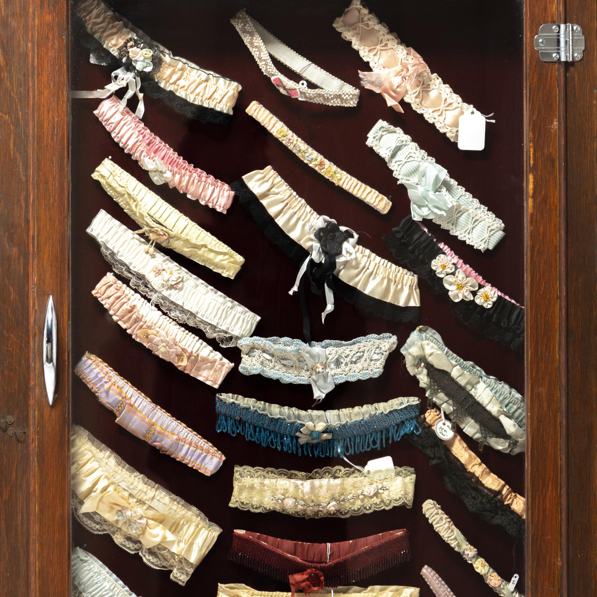 Fantastica esposizione a vita di giarrettiere da salone/bordello da donna in un'elegante vetrina di Oak. Pizzo fantasia, seta, fiori, perline, perline a telaio, tema western, etc. per un totale di 79 pezzi. Ottime condizioni. Un'esposizione
