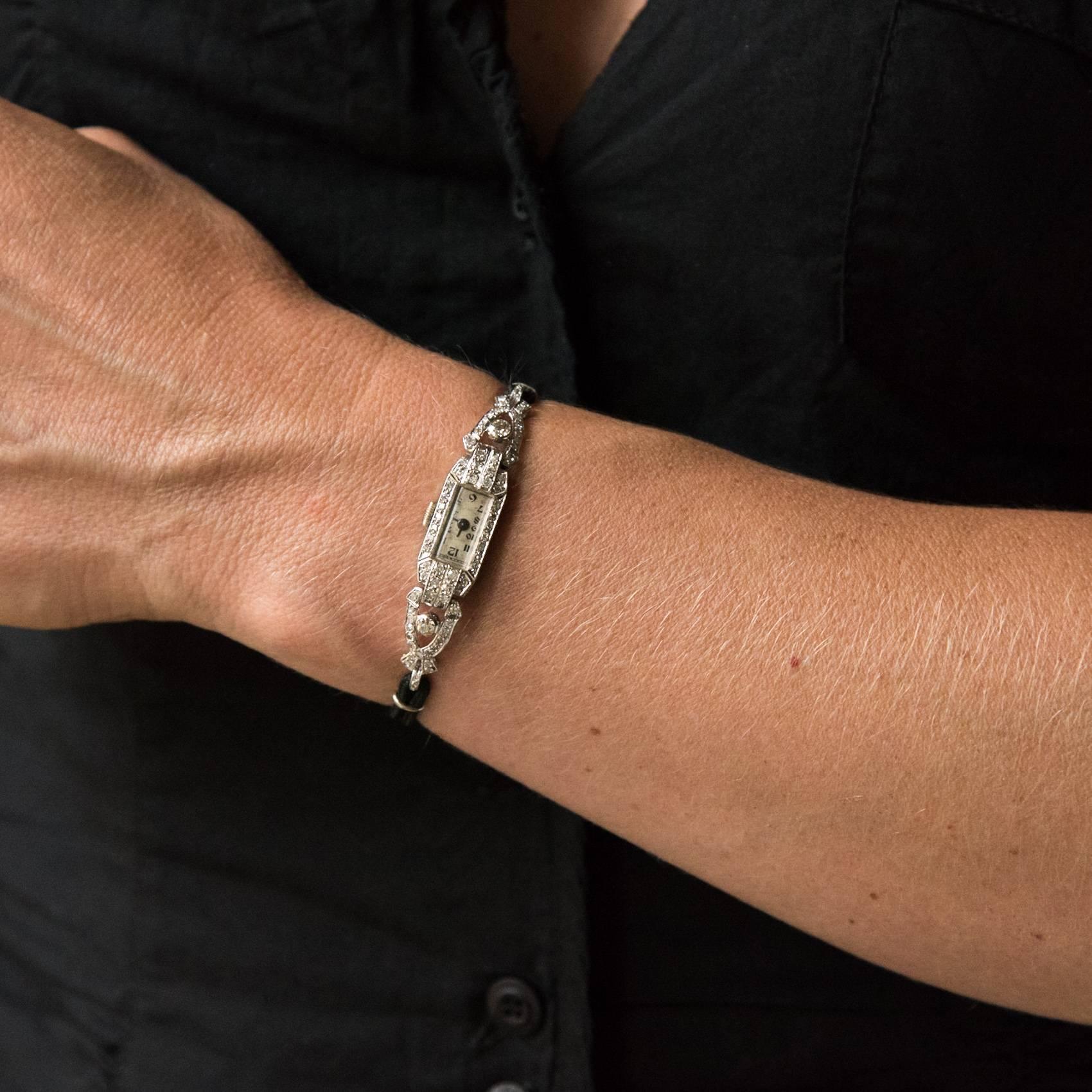 Rectangulaire, cette montre art déco présente des lignes géométriques et des décorations serties de diamants.
Le bracelet est un double cordon noir retenu par des liens en or blanc. Le fermoir en or blanc est une boucle déployante avec cliquet et