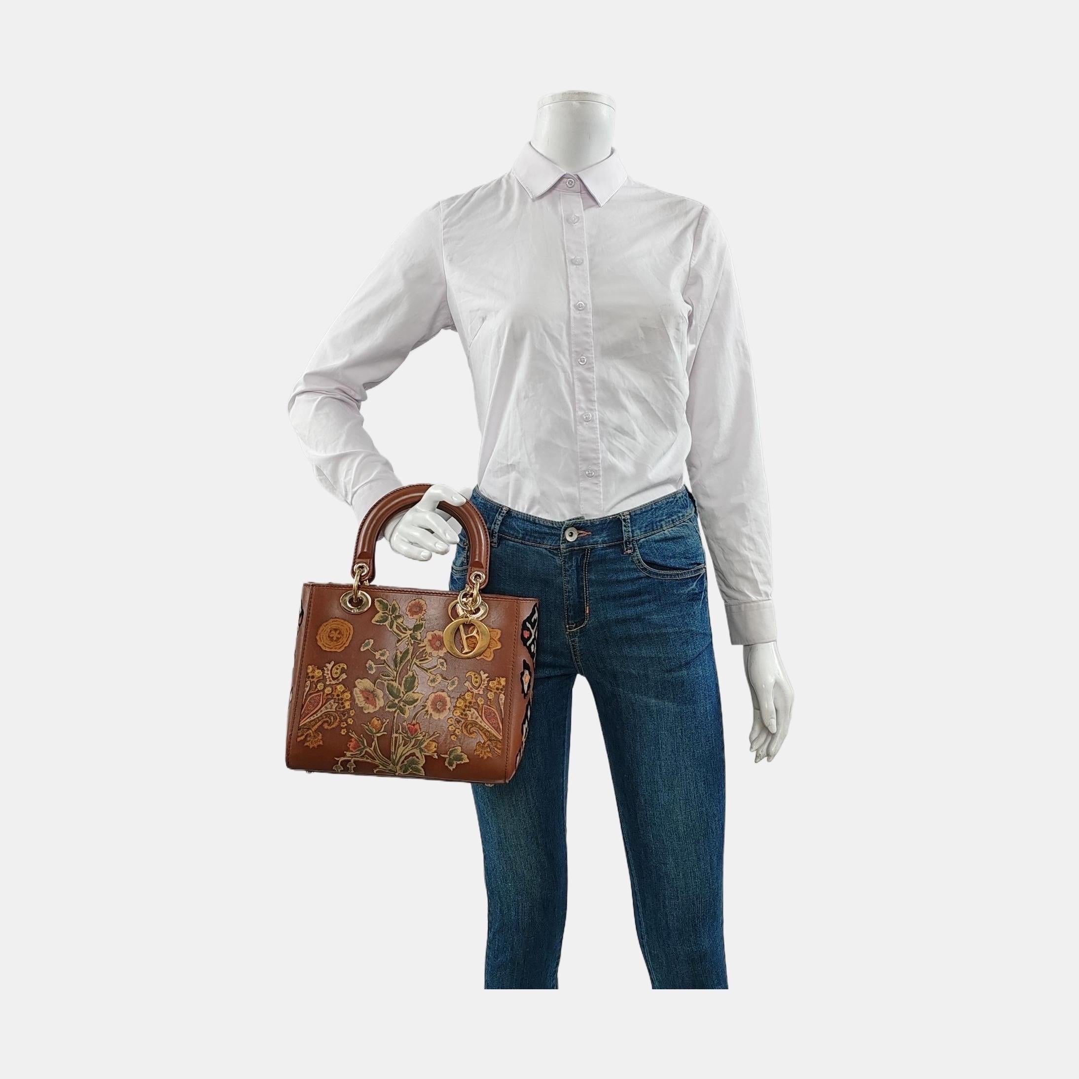 This tote showcases a laser-cut floral design on rich brown calfskin leather. It is equipped with durable leather top handles and accented by gold hardware, including signature D.I.O.R. initial charms. The top opens to reveal a leather-lined