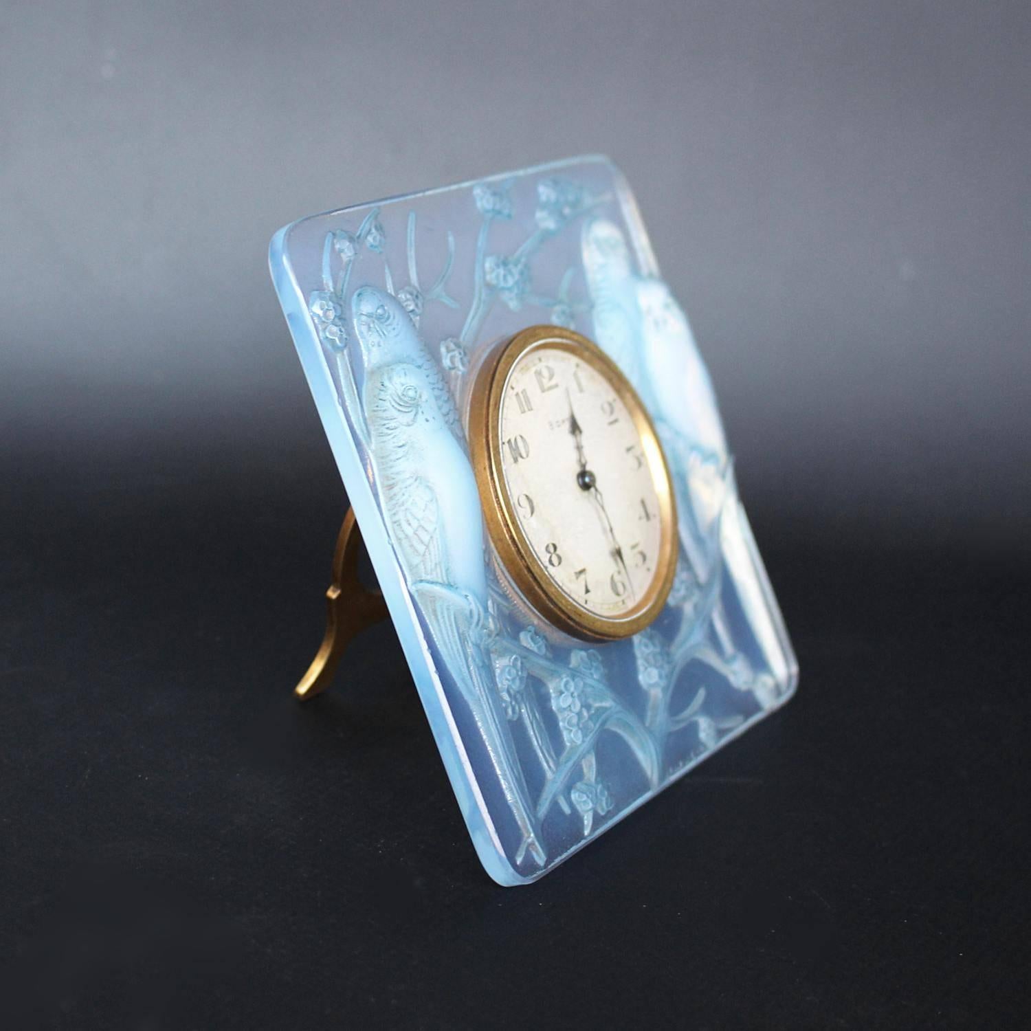 Inséparables, an Art Deco glass clock. Moulded in relief with two pairs of frosted lovebirds with original turquoise highlights, seated on prunus blossom. Cream coloured clock face. Eight day Swiss movement and hinged stand.

Raised R Lalique to