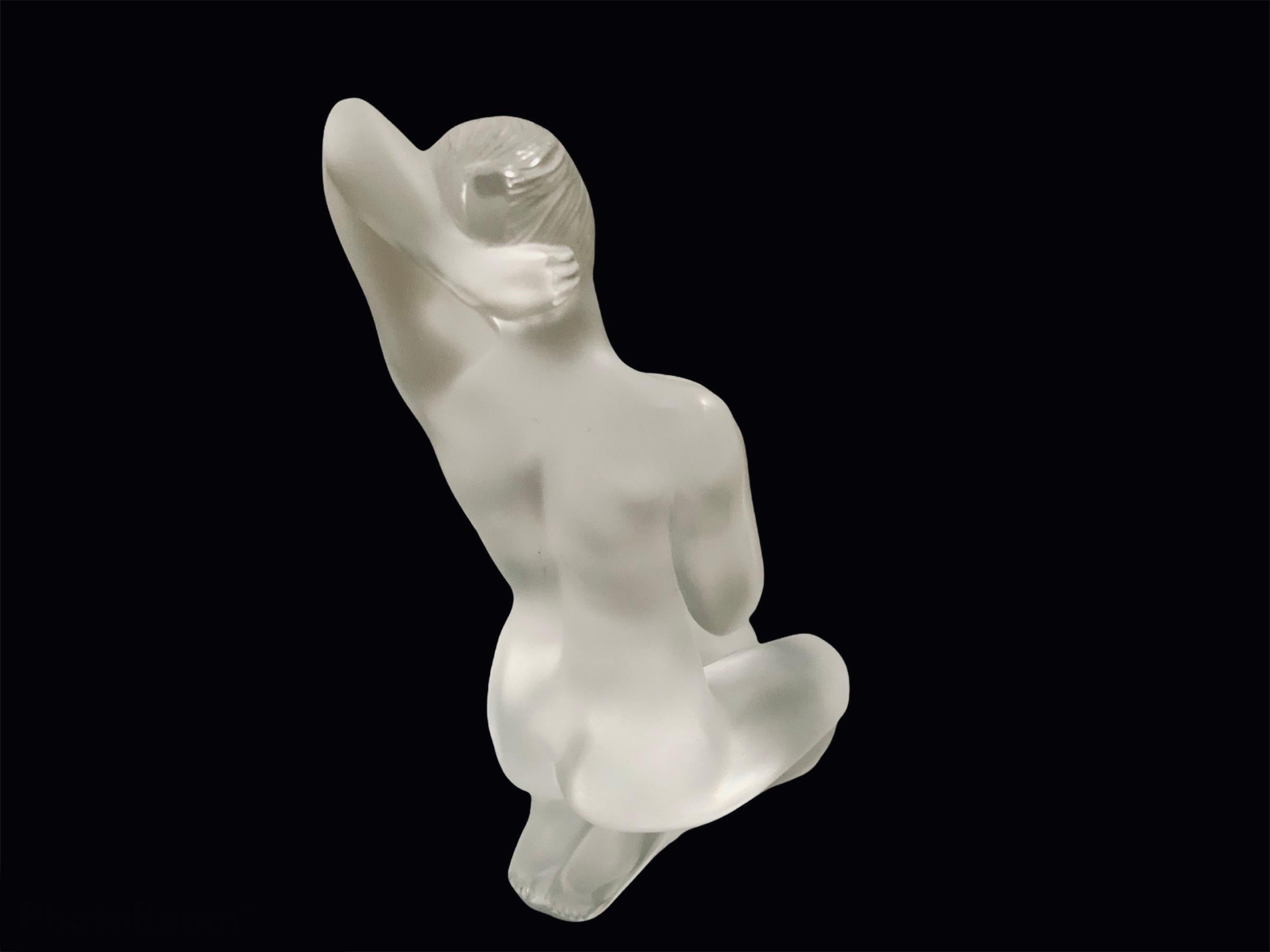 This is a Lalique clear crystal small aphrodite nude sculpture. It depicts a nude woman in a sensual kneeling position with her right hand over the same side shoulder and the left one behind her head. The sculpture is acid etched signed