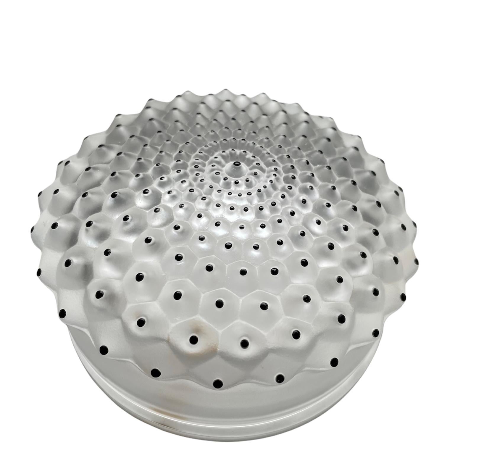 This is a Lalique Crystal Cactus vanity powder box. It depicts a round frosted crystal with a concave lid. Over it, you can see a cluster of frosted crystal lumps ending with black enamel dots. There is a Lalique France sticker in one of the side