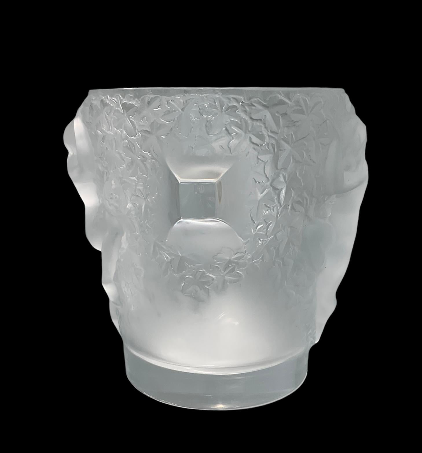 This is a Lalique crystal Ganymede champagne cooler/ice bucket. It is a wide, heavy and cylindrical shaped clear and frosted crystal container. It is decorated by bas-reliefs of nude dancers in a background of grapes leaves. In Greek and Roman