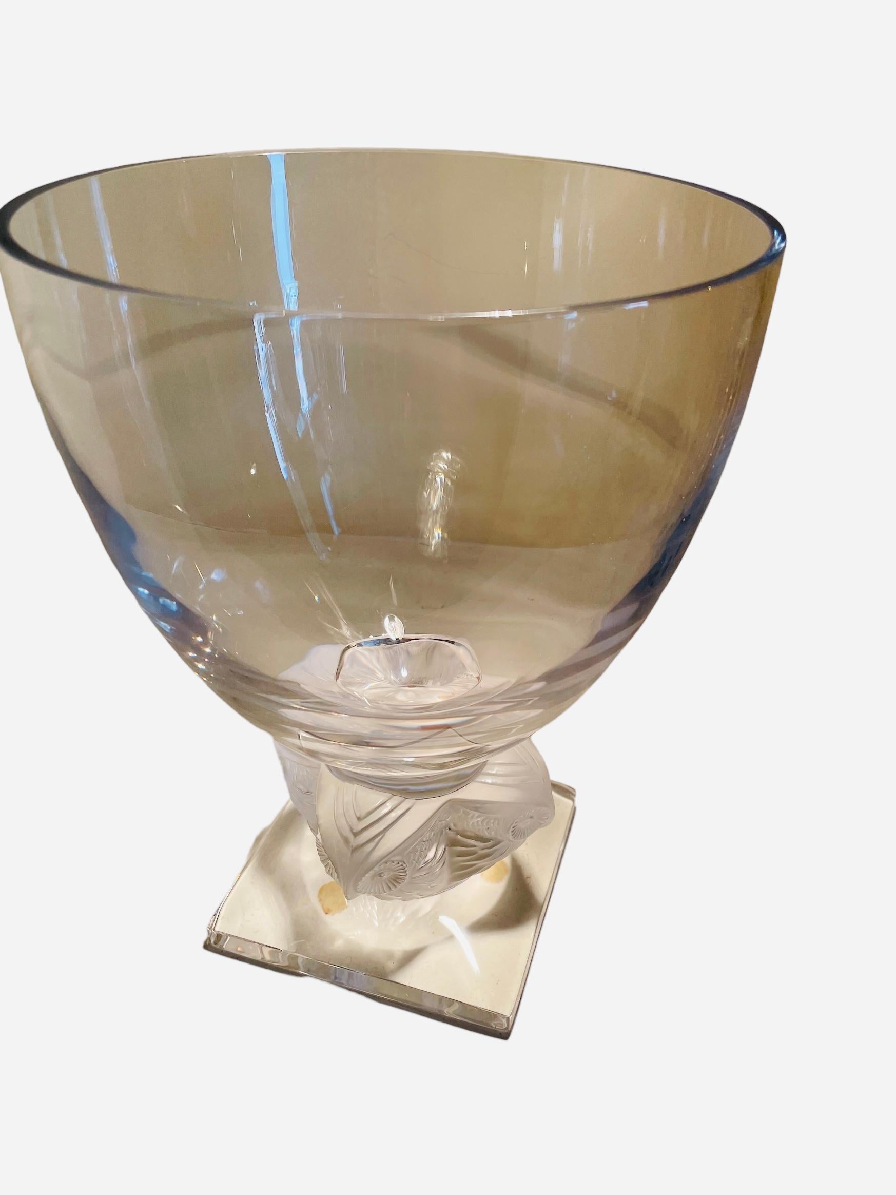 This is a Lalique Crystal “Grand Ducs” Owl flower vase. It depicts a large round bowl that is supported by the frosted crystal heads of four owls. Then, they are attached to a clear crystal square base. At one of the border of the base, it is found