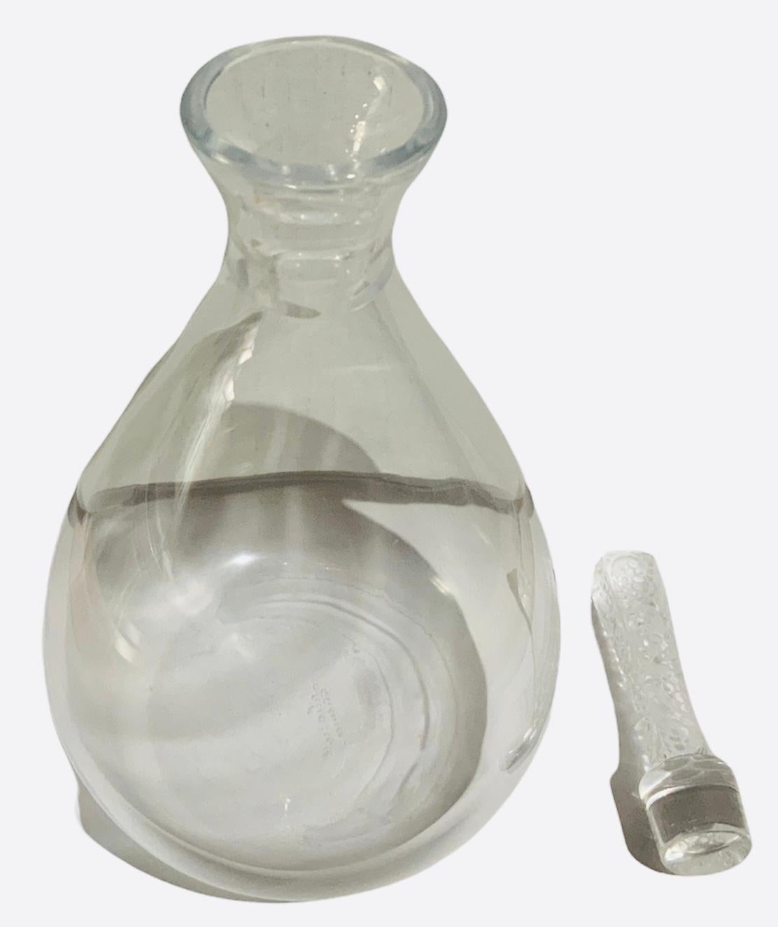 This is a Lalique Crystal Phalsbourg decanter. It depicts an urn shaped clear crystal bottle with a long tapered-rectangular stopper. The stopper is adorned with an etched pattern of long scrolls with grapes. Below the bottle, it is found the