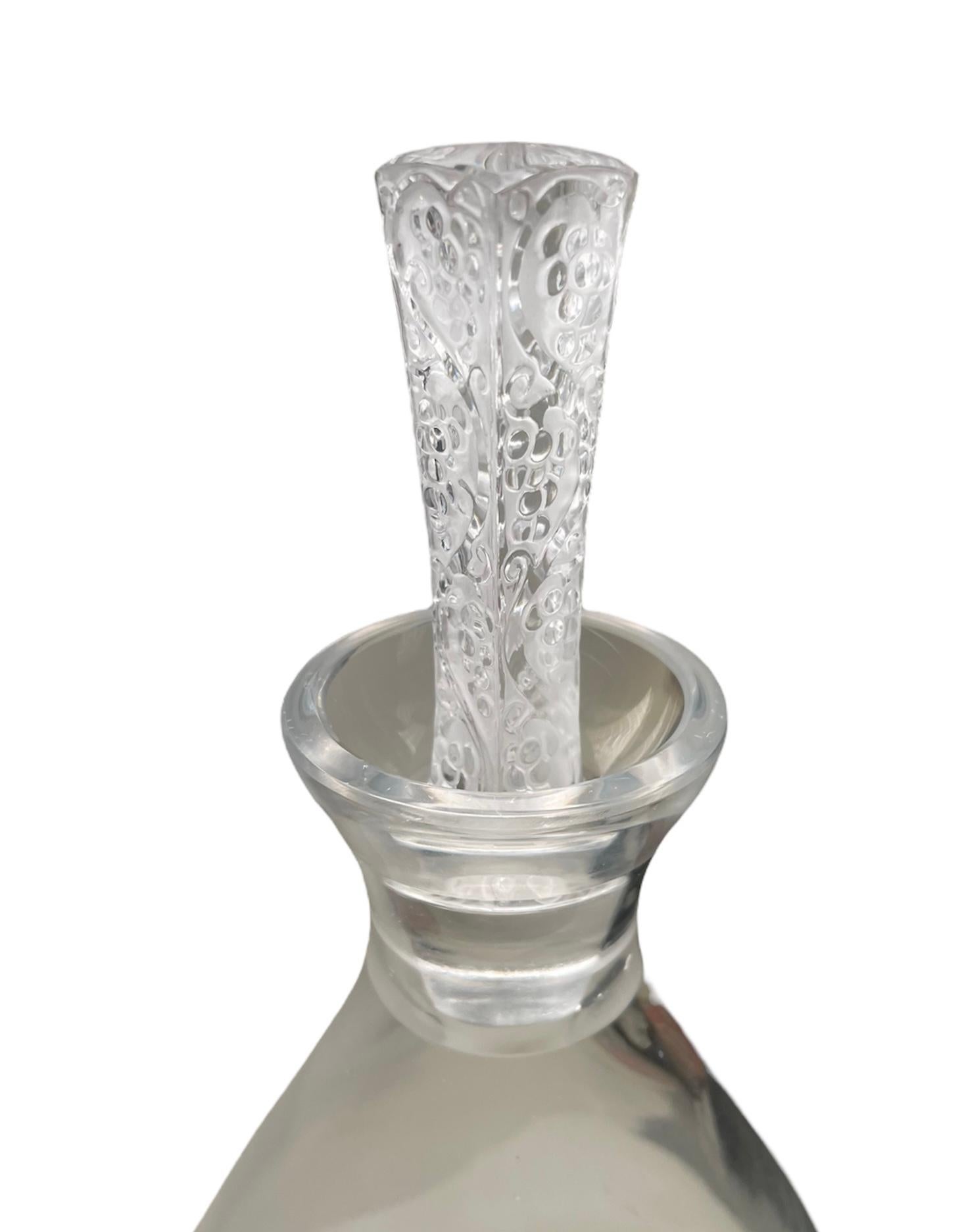 This is a Lalique Crystal Phalsbourg decanter. It depicts an urn shaped clear crystal bottle with a long tapered-rectangular stopper. The stopper is adorned with an etched pattern of long scrolls with grapes. Below the bottle, it is found the