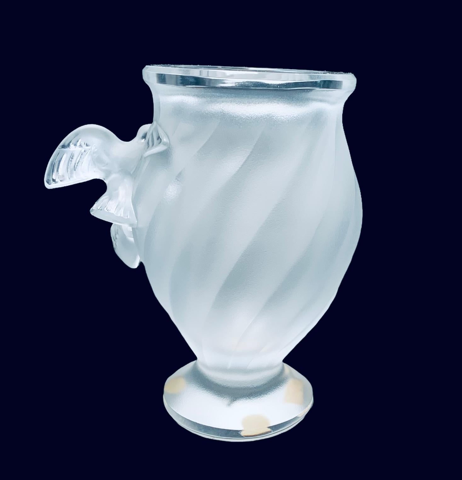This is a Lalique clear/frosted crystal vase. It depicts two Rosine birds on flight in the center of the vase. There are some delicate ribbed swirls adorning the crystal. The Lalique, Paris sticker is under the base.