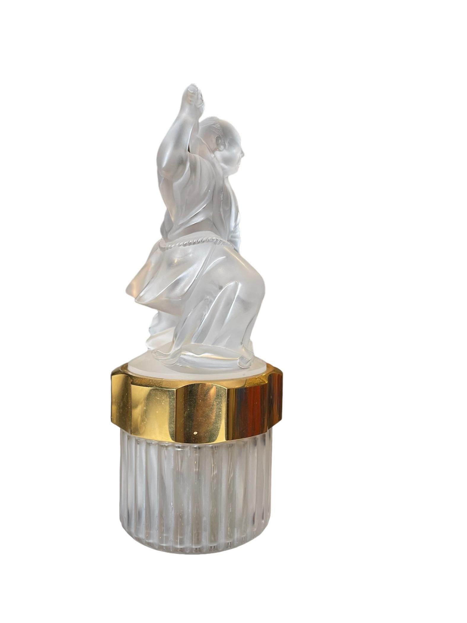 This is a Lalique Crystal Sculpture Perfume Bottle. It depicts a clear and frosted crystal Samurai sculpture standing over a round crystal bottle that is covered by a gold tone plastic round lid. The clear and frosted cylindrical bottle is ribbed.