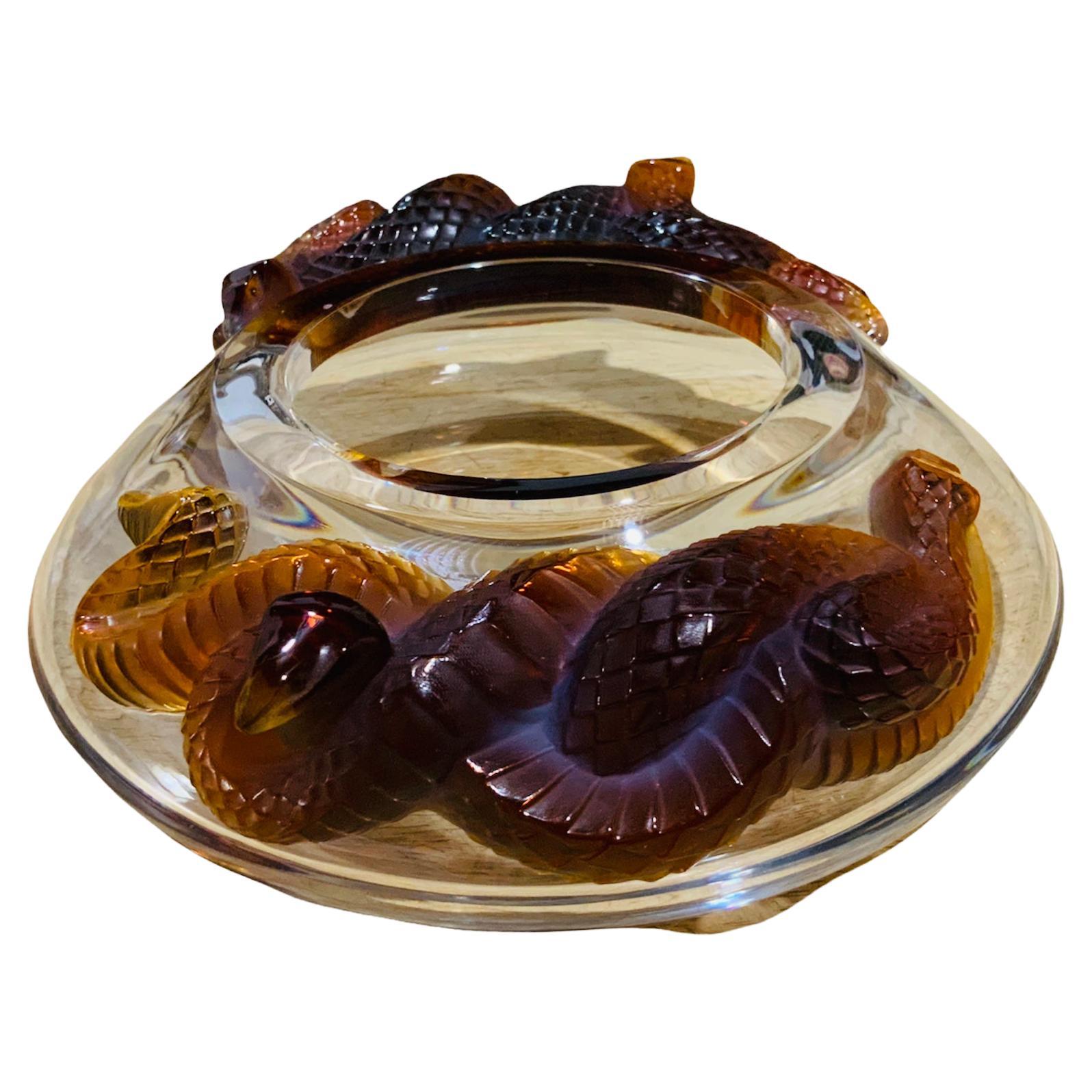 This is a Lalique, France Serpent bowl. It depicts two amber-brown color crystal snakes intertwined at each side of a wide round clear crystal bowl. Below the base, it is the acid etched hallmark of Lalique. Snakes symbolize intelligence, wisdom,