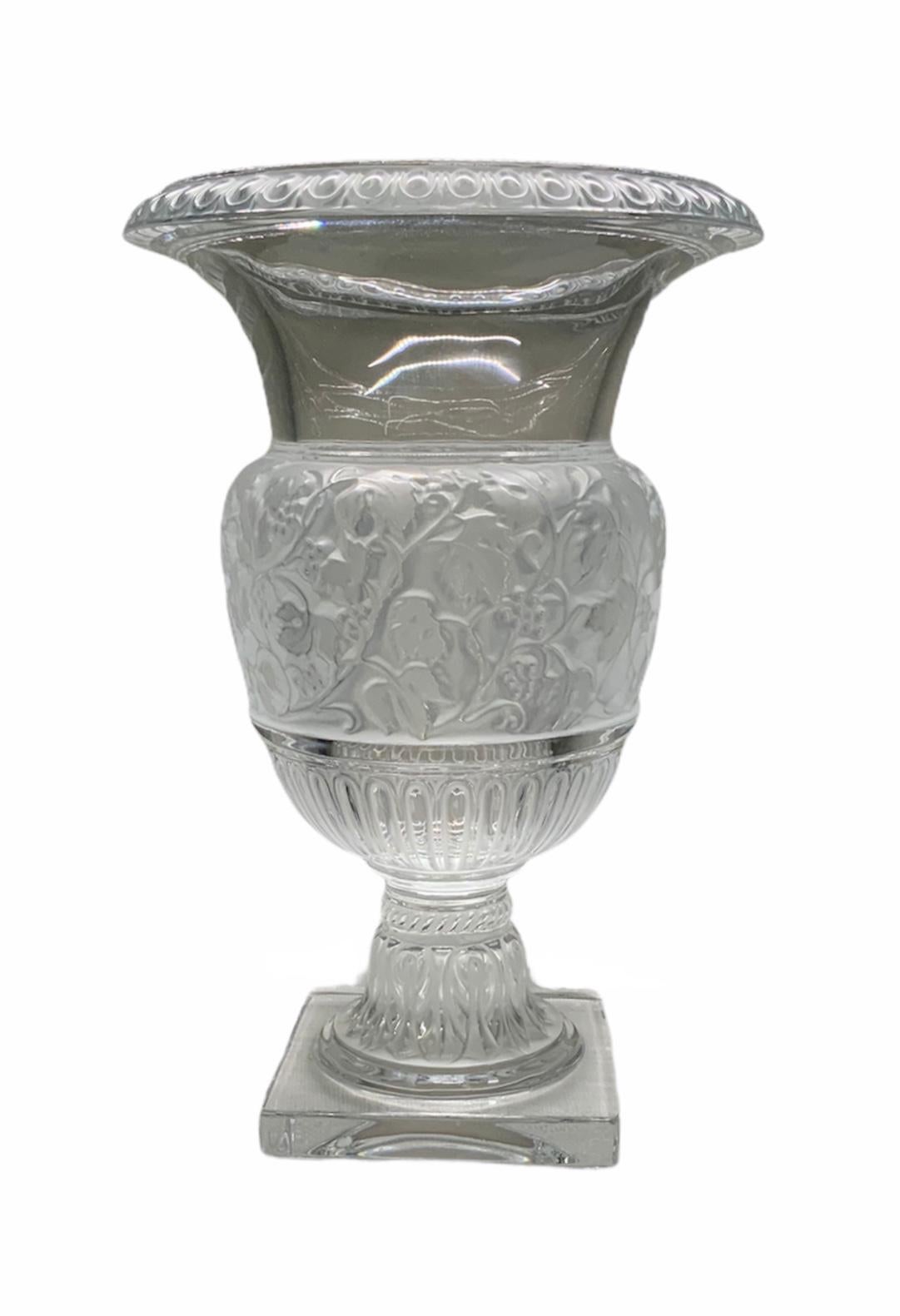 This a large clear Lalique crystal urn vase adorned with a continuous carved flowers branches. The vase has a bulbous body and a bell shaped rim. It is signed Lalique in the border of the square base.