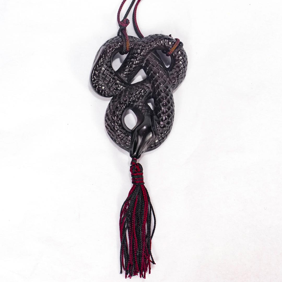 A fine French art glass pendant necklace.

By Lalique.

Entitled: Sautoir Serpent Noir.

Comprised of a black coiled Lalique glass snake with a black & burgundy tassel in its mouth. 

Strung on 2 black and burgundy cords.

Signed Lalique to the