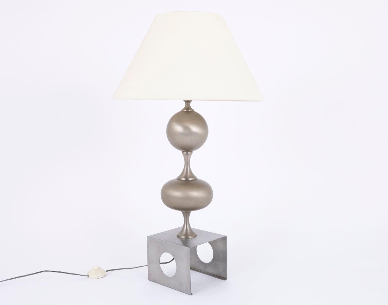 Lamp in brushed stainless steel with a shaft composed by a round ball and a flatened ball, linked each other by small stylized hourglasses. Square shape base with three sides, drilled with two holes on each side of the lamp.

Work realized in the