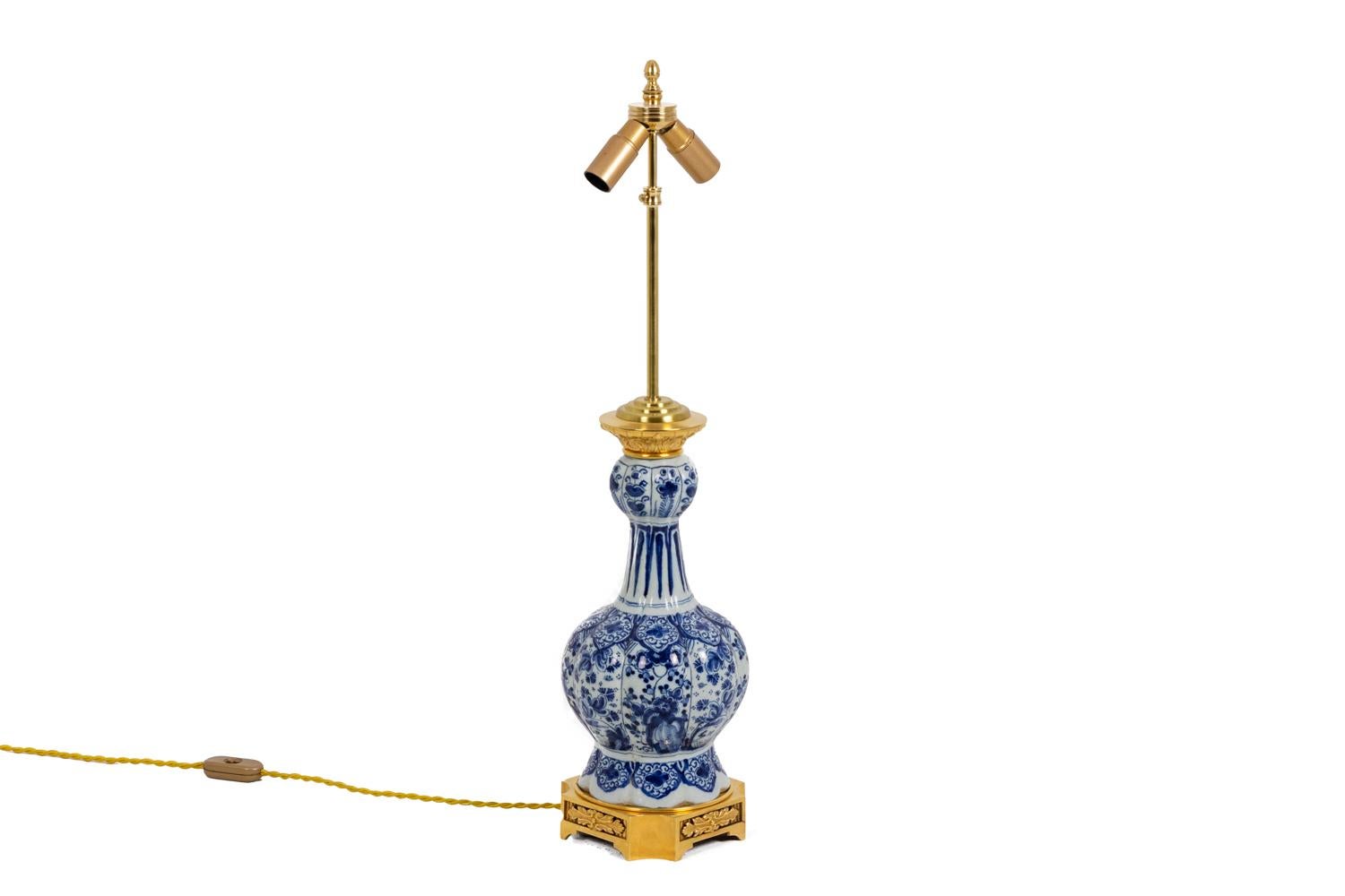 Baluster shape lamp in blue and white Delft earthenware adorned with flowers and foliages Paunch of gadroons supporting a ball adorned with flowers. Mount in chiseled and gilt bronze. Quadripod cut base and adorned with foliages in grooved bottom