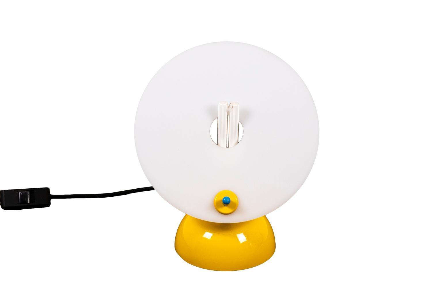 Lampe en Lucite composée d'une base en demi-sphère jaune supportant une plaque circulaire inclinée blanche percée en son centre. Interrupteur bleu de montée et descente.

Travail réalisé dans les années 1970.

Le collectif Memphis a été créé à