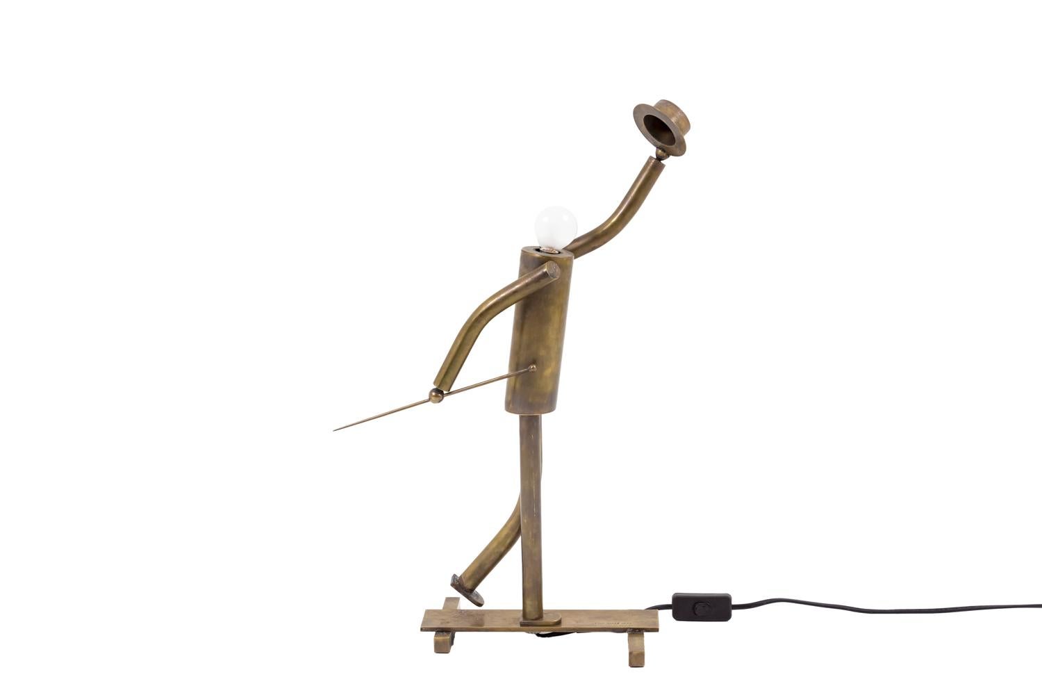 Lamp in patinated brass figuring a stylized music-hall dancer holding a hat in his raising hand and a stick with a round knob in the other hand. The bulb represents the character head. He raises his back leg and the other one is standing on a