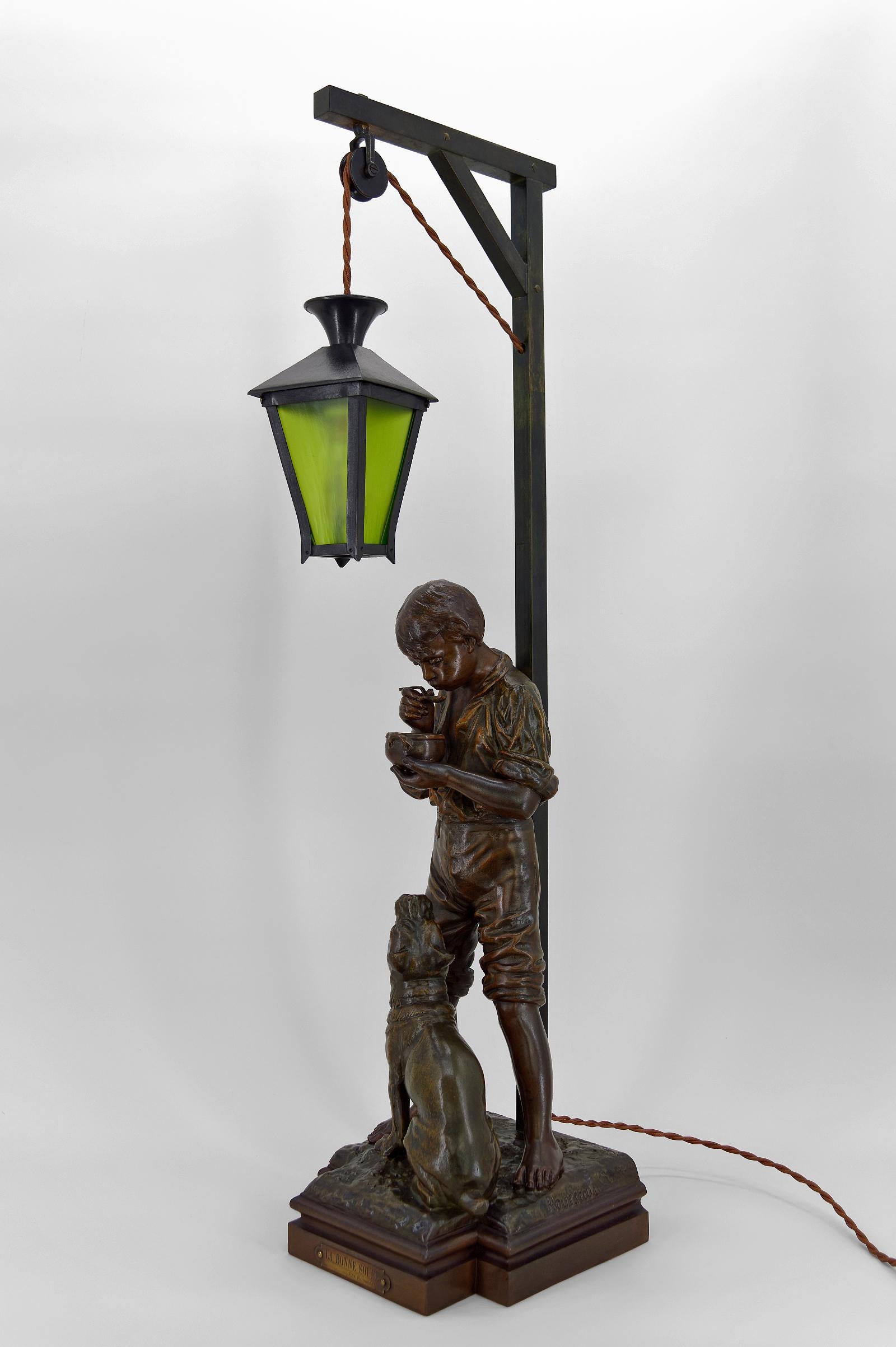 Important patinated spelter lamp representing a young boy, probably a butcher boy, drinking his soup accompanied by his pit bull / bulldog type dog.
The boy is leaning against a gallows holding an electrified green lantern.

By Belgian sculptor